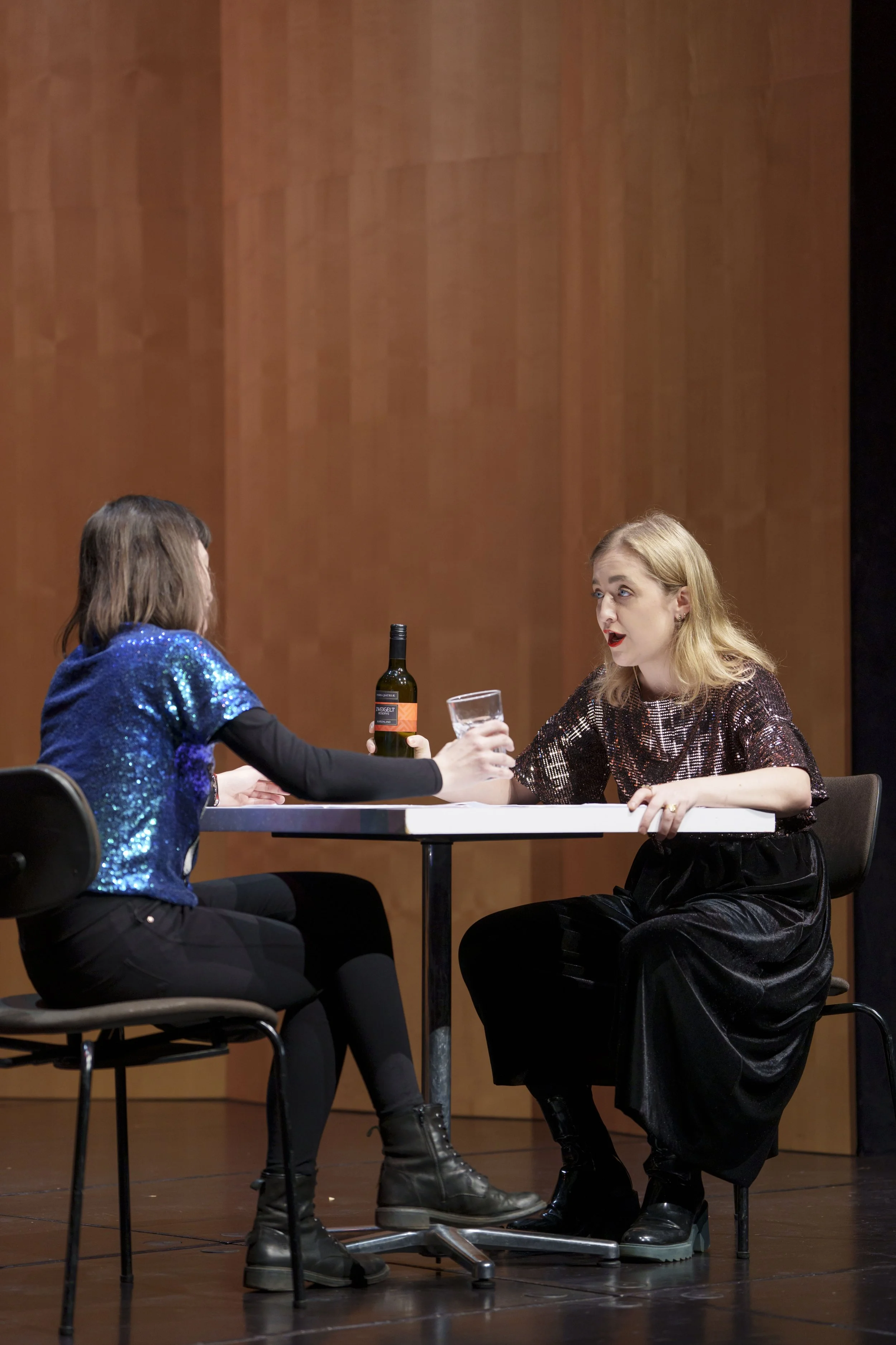 Two women are sitting at a table on a stage. One woman has brown hair and is wearing a blue sequined top, and the other has blonde hair and is wearing a shimmery top. They are engaging in a conversation, with the blonde woman holding a glass and the 