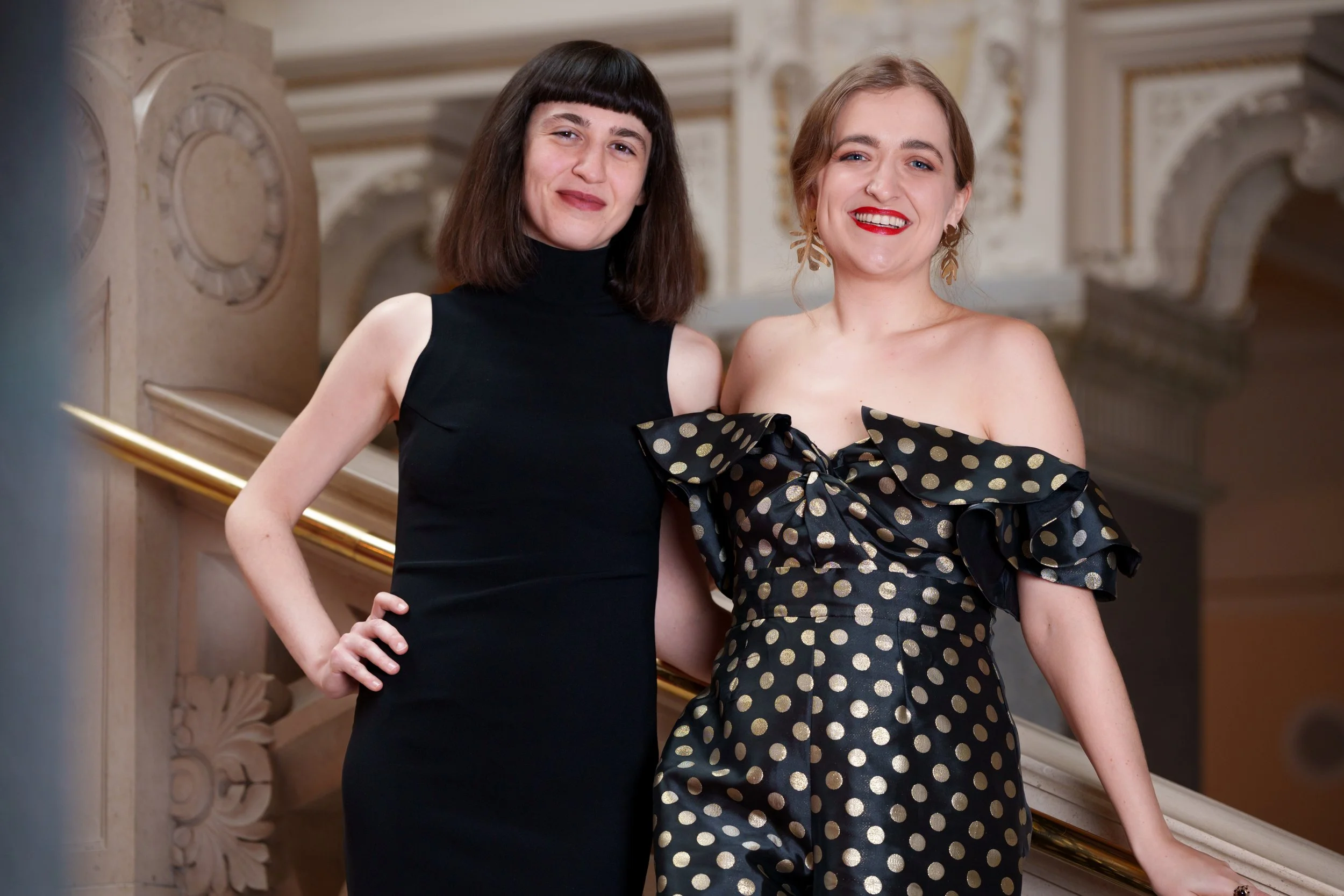Two women standing on a staircase, smiling at the camera, dressed in elegant black and gold formal attire, with an ornate architectural background.