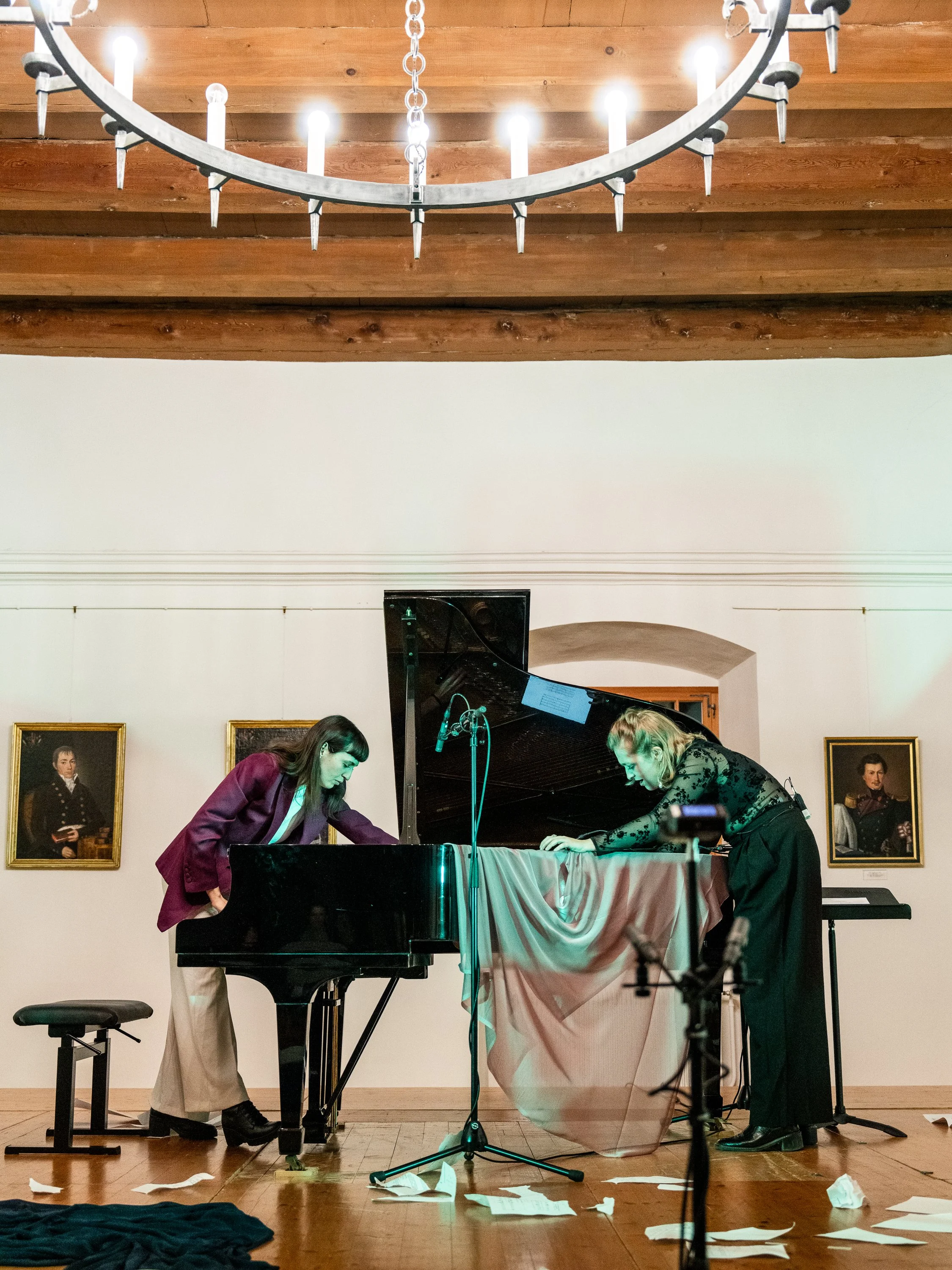 Two women are bowing after a musical performance with a grand piano in a room with portraits on the wall and a chandelier hanging from a wooden ceiling.