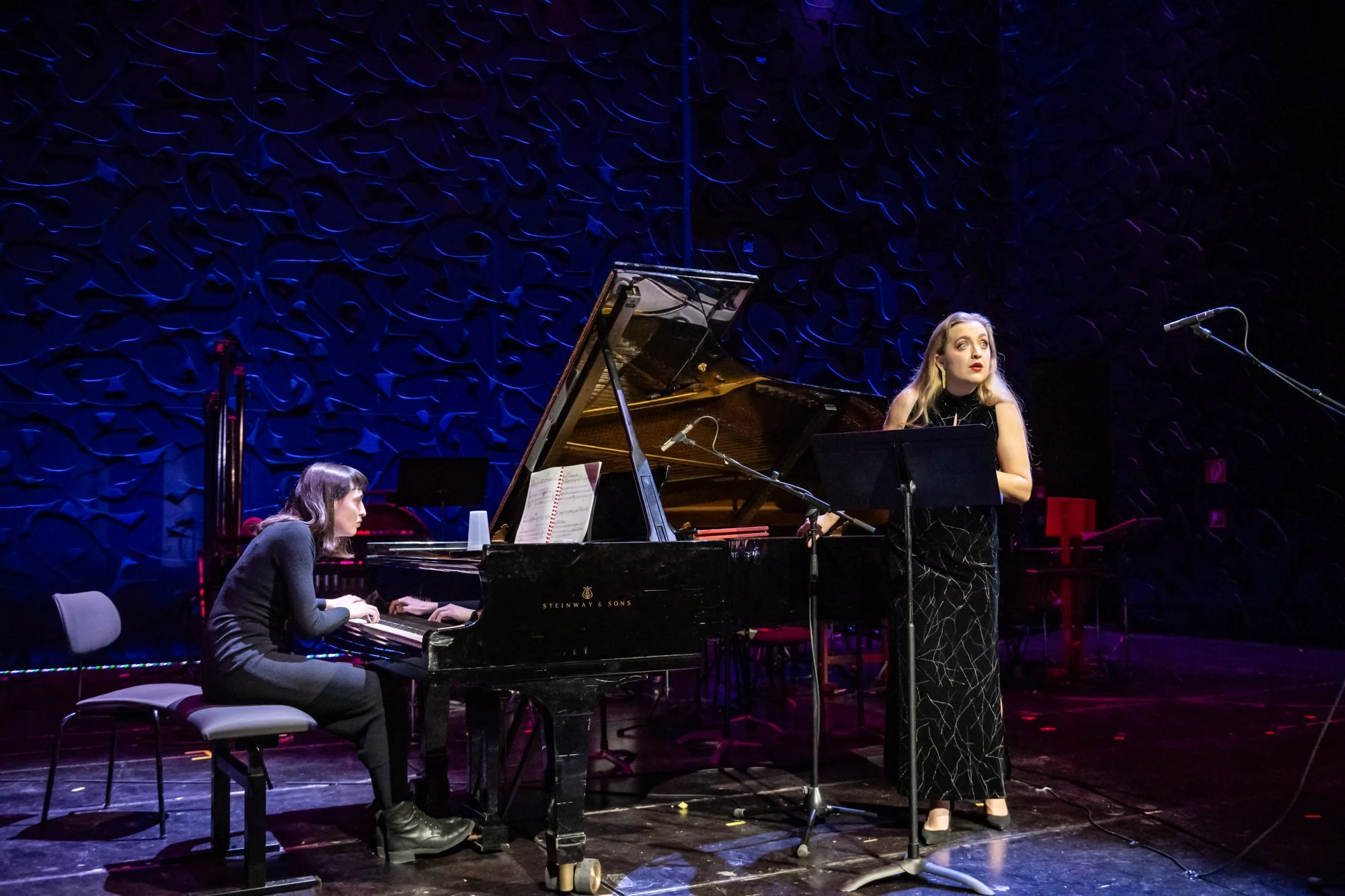 Two women performing jazz music on stage, one playing the piano and the other singing in front of a microphone, with blue textured back wall.