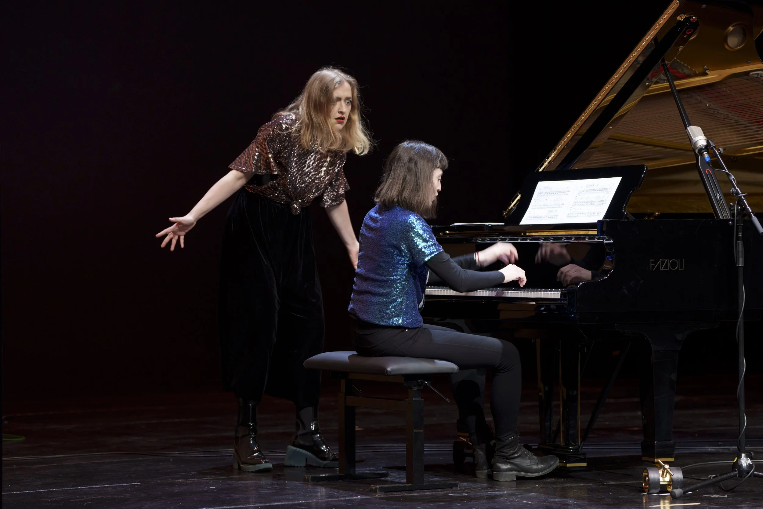 Two women, one sitting and playing piano and the other standing and singing, on a dark stage. The pianist is wearing a blue sequined top and black pants, and the singer is in a shiny dark shirt and black boots.