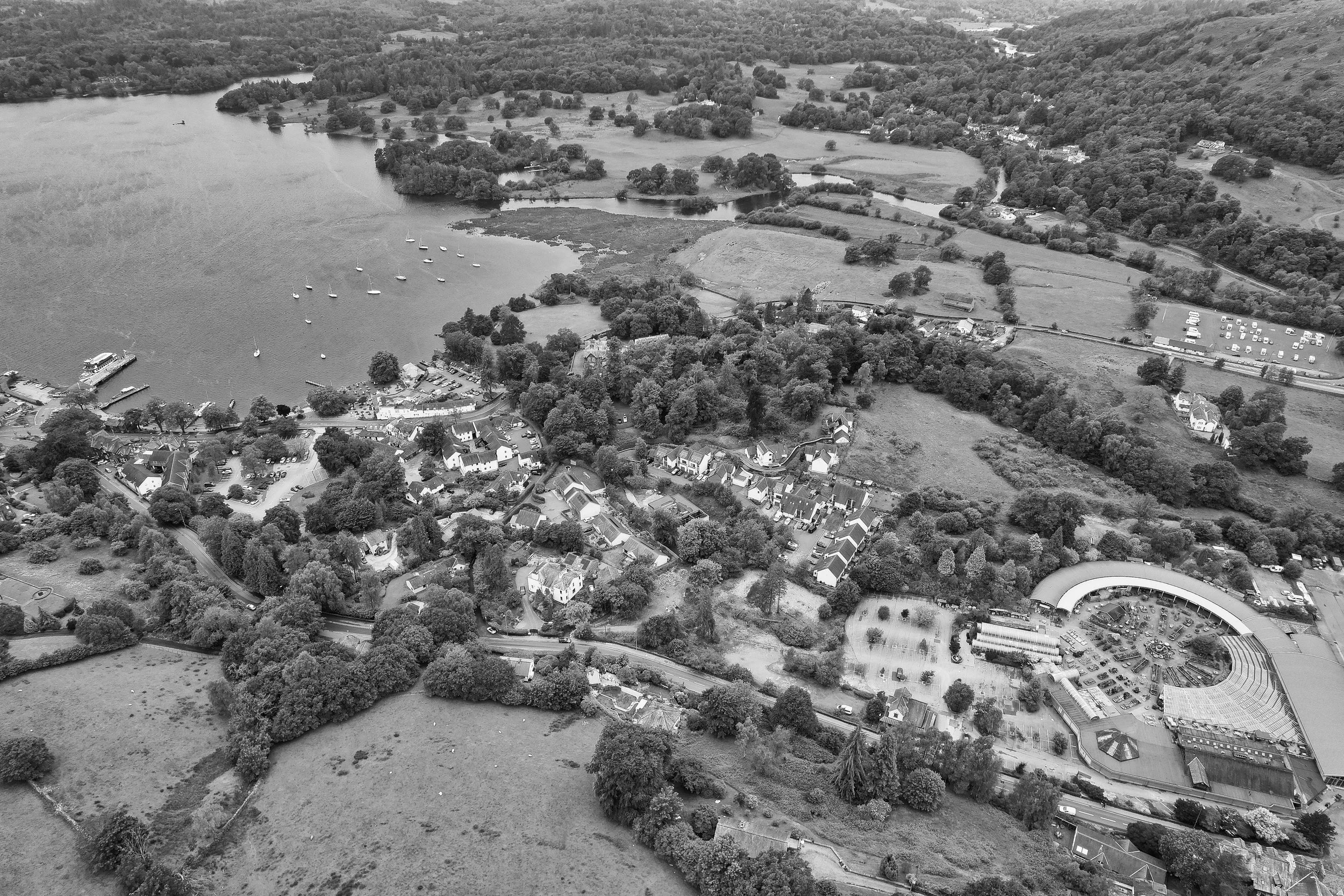 Aerial view of a lakeside village surrounded by trees, with boats docked at the shore, residential houses, a park, and a curved building in a rural area.
