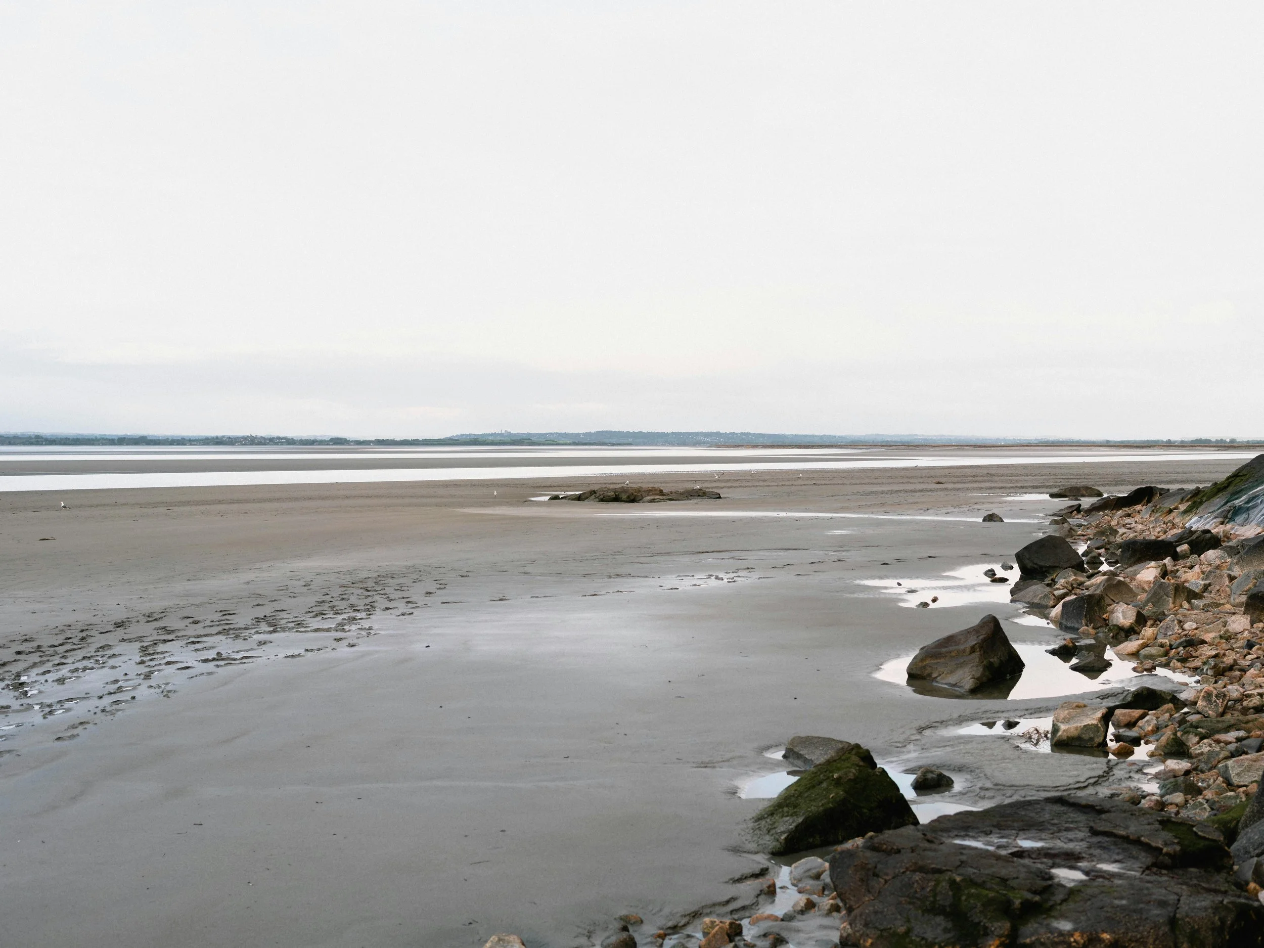 A wide view of a beach with mostly wet sand, some rocks along the shoreline, and a stormy, overcast sky.