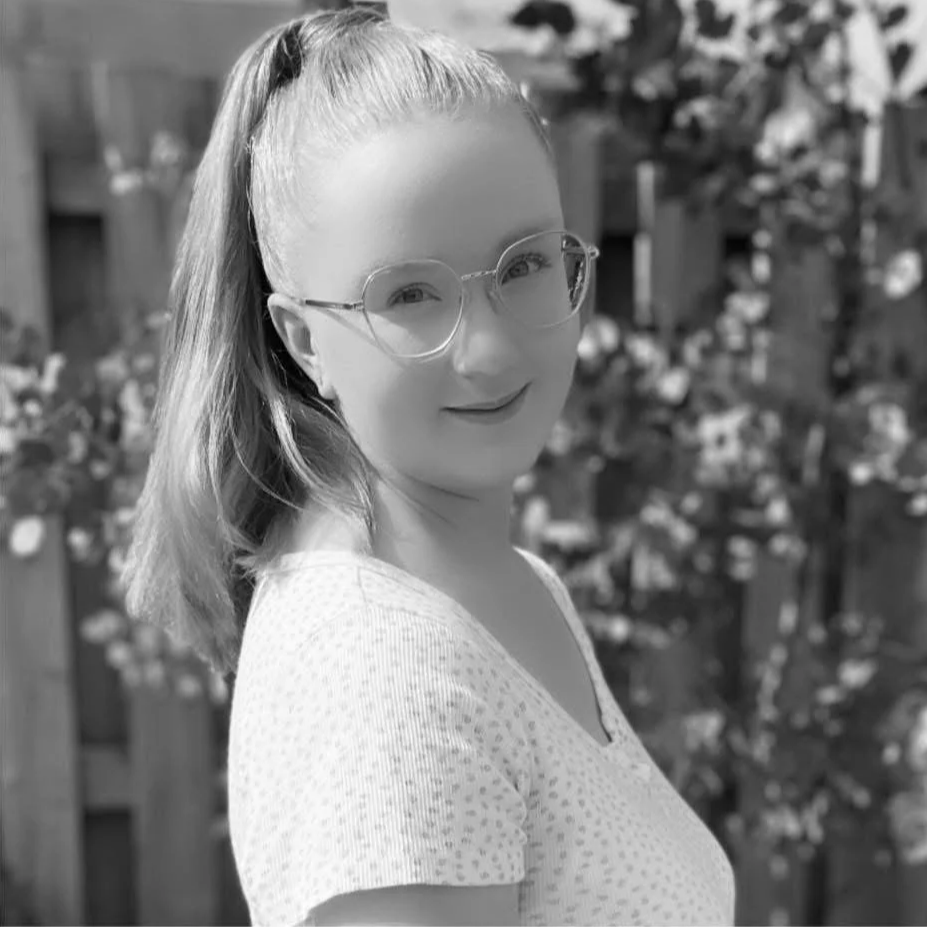 A young woman with long hair tied back, wearing large round glasses and a light patterned shirt, smiling outdoors with foliage in the background.