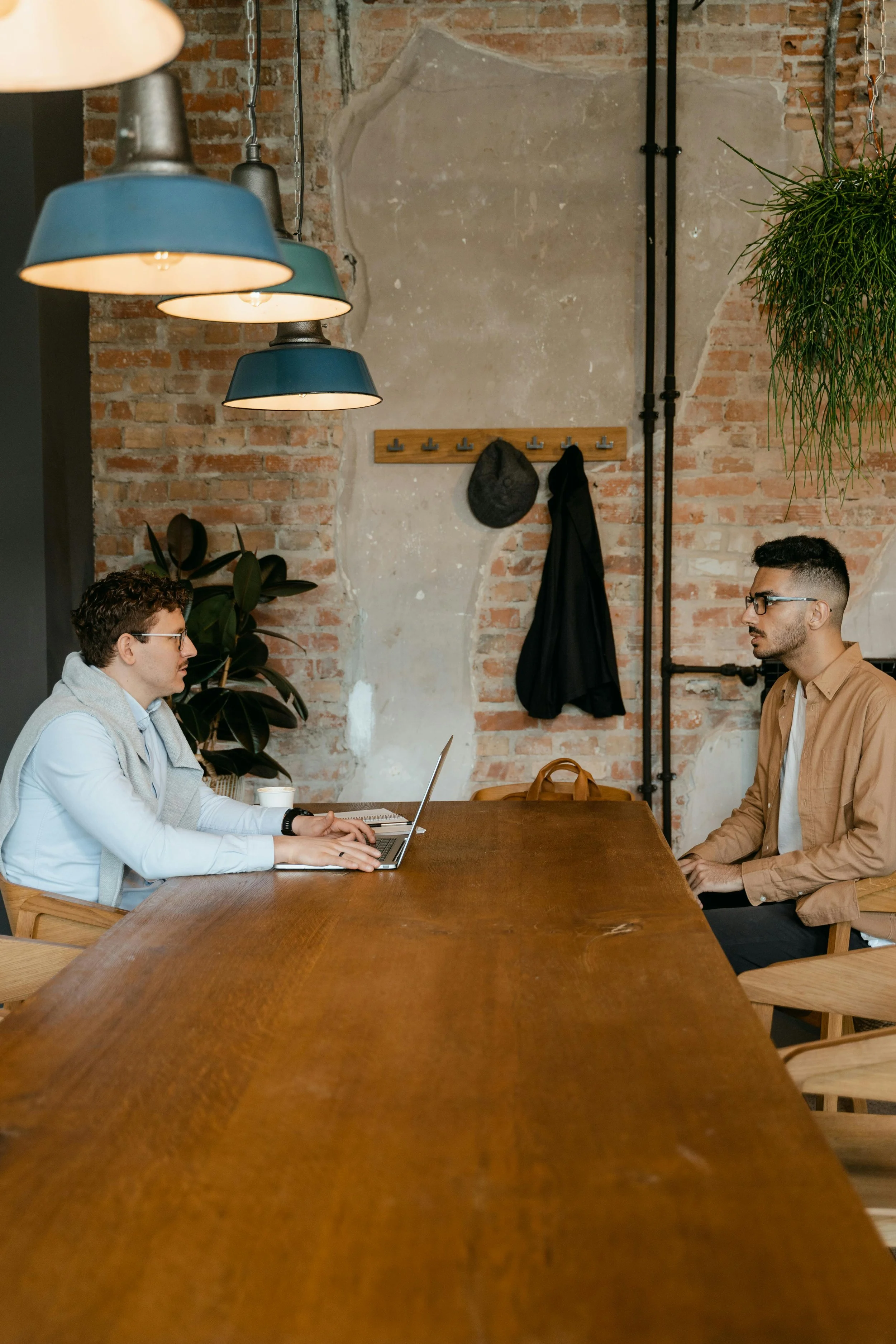 Two men sitting at a long wooden table in a modern, industrial-style room, facing each other, with a brick wall and hanging lamps above.