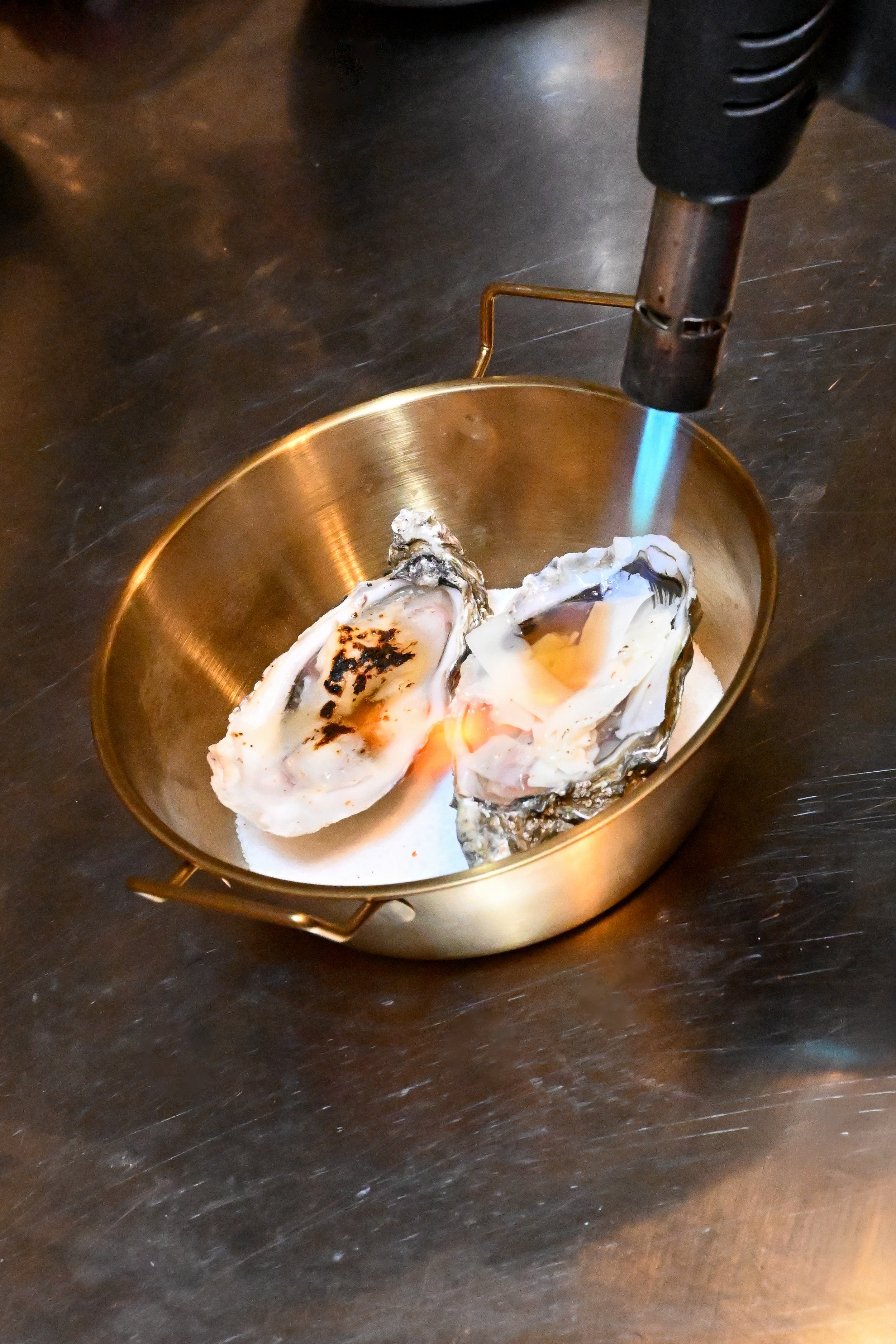 Deux huîtres ouvertes dans un plat en métal, avec un chalumeau torche à proximité sur une surface en bois sombre. Une entrée proposée par la cheffe avec des produits de qualité et de saison.