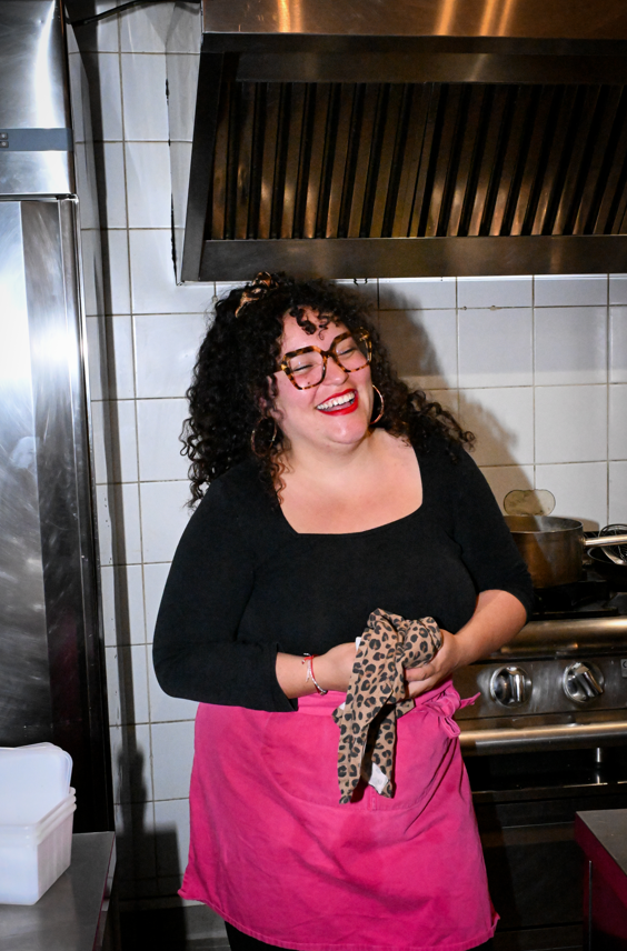 Femme souriante portant un tablier rose, tenant une serviette avec un motif léopard, dans une cuisine. Alizée Rat cheffe du restaurant la Baignoire by Lili.