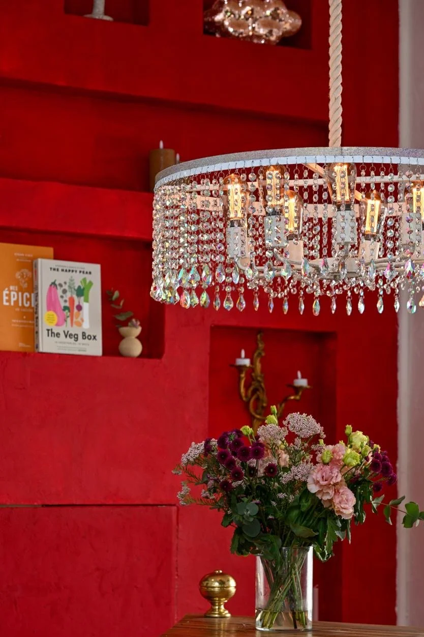 Un lustre en cristal suspendu avec une corde, au-dessus d'un vase de fleurs aux couleurs roses, blanches et violettes, dans une pièce au mur rouge avec des étagères et des bougies. Une ambiance cosy idéale pour un restaurant en amoureux, en famille.