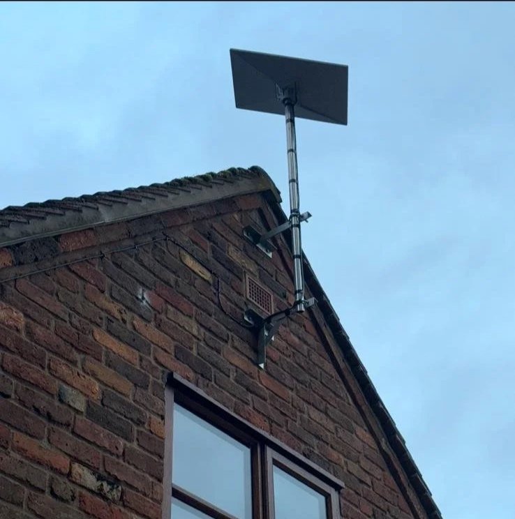 A Starlink dish installed on the roof of a brick house, pointing upwards against a cloudy sky.