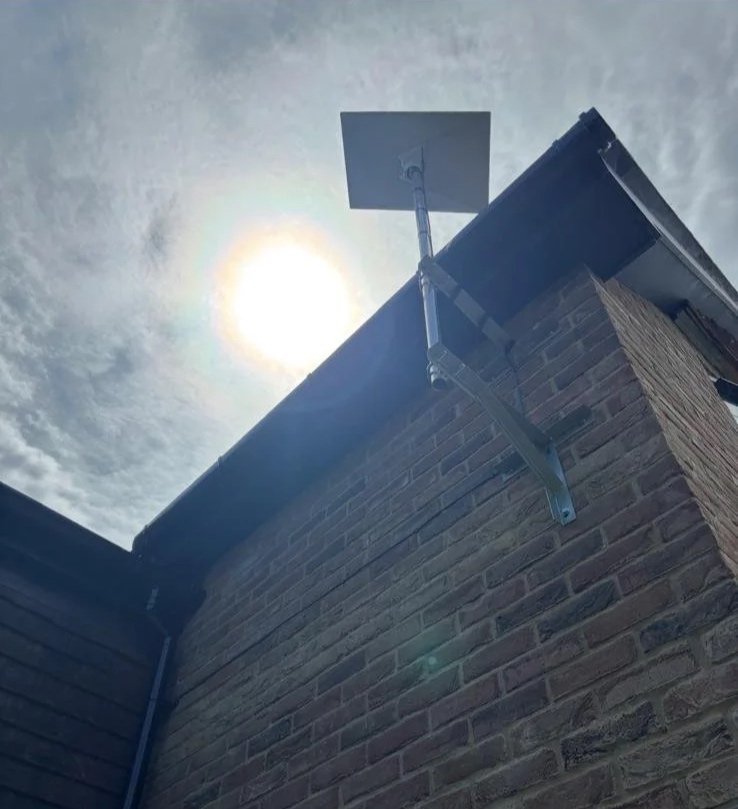 A Starlink dish mounted on a metal pole on the corner of a brick building with the sun shining in the sky.