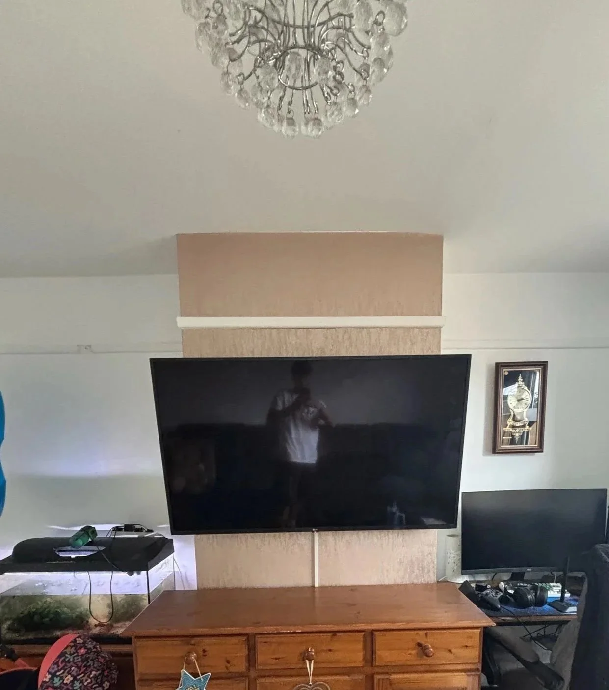 Living room with a wall-mounted, blank black flat-screen TV above a wooden cabinet, a chandelier on the ceiling, and a framed picture on the wall to the right of the TV.