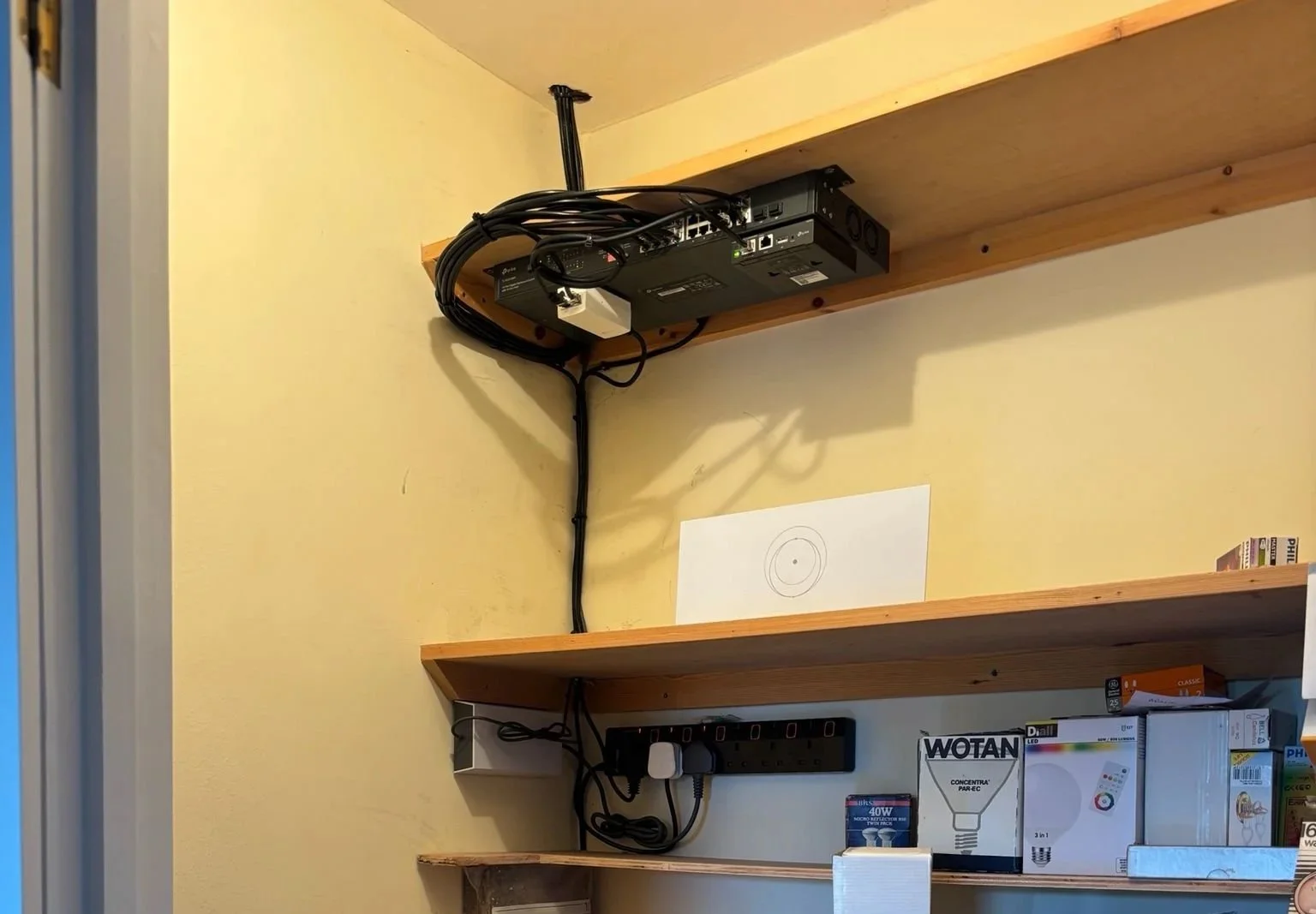 Two wooden shelves mounted on a yellow wall, with electronic equipment and boxes on the lower shelf. The upper shelf holds a black electronic device with multiple cables connected, mounted on a wooden board. A white paper with a circular diagram is o