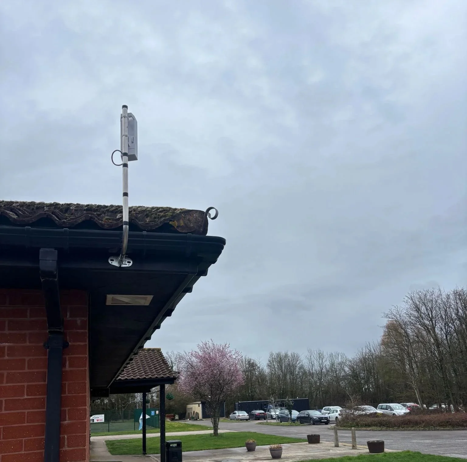 Overcast sky above a parking lot with some trees and a brick building with a black roof. An antenna is mounted on the roof.