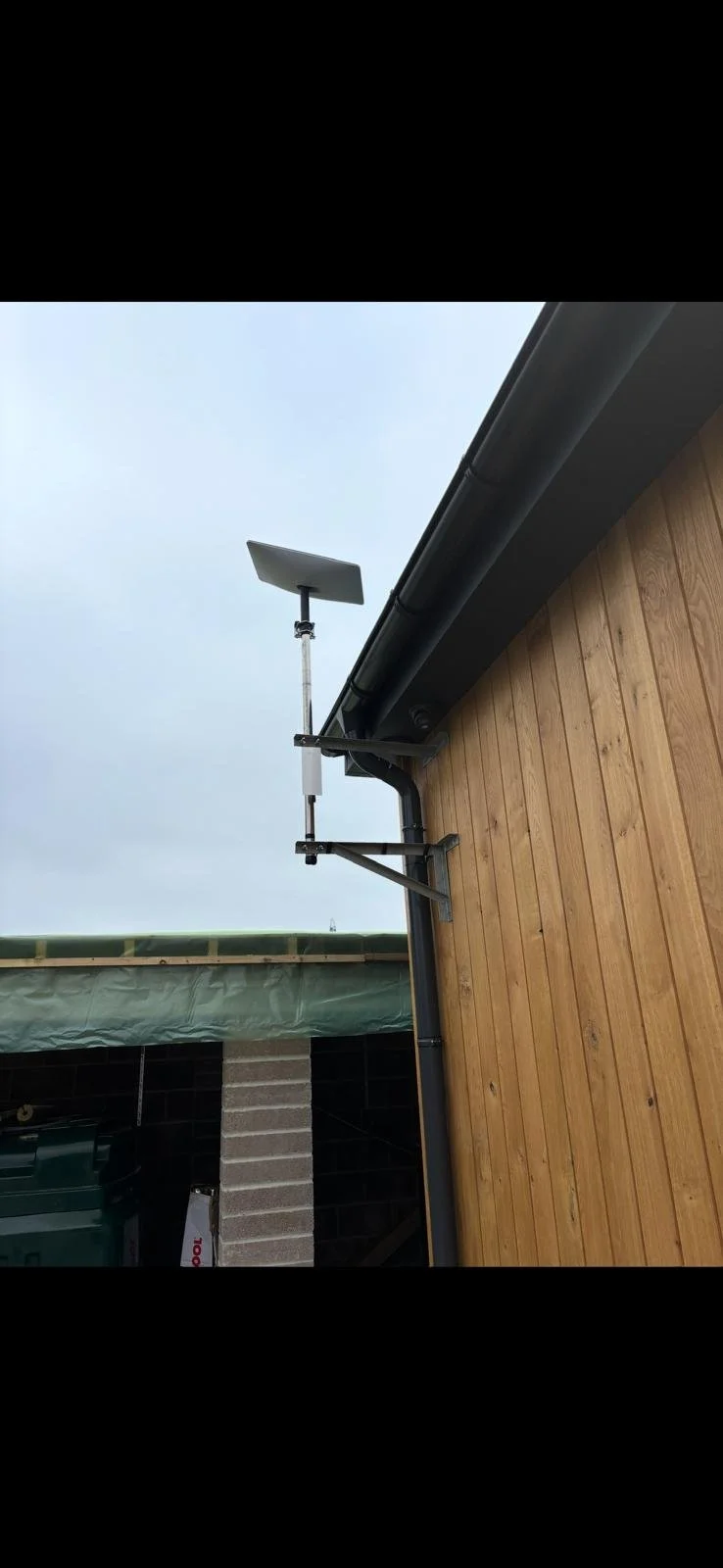 A Starlink dish mounted on the side of a wooden building with a black gutter, during daytime with a cloudy sky.