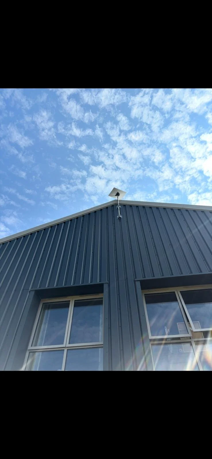 View of the corner of a building with dark metal siding and two windows, looking up at a blue sky with scattered white clouds, and a small antenna mounted on the roof.