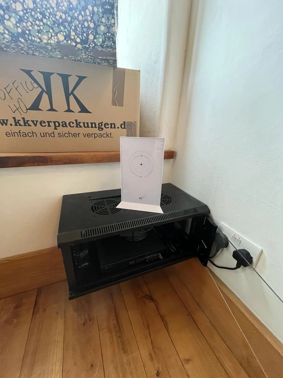 An electronic device with a circular patterned design on top, placed on a wooden floor next to a wall socket with a plugged-in power cord. A sheet of paper with a target diagram is leaning against a small shelf or packaging in the background.