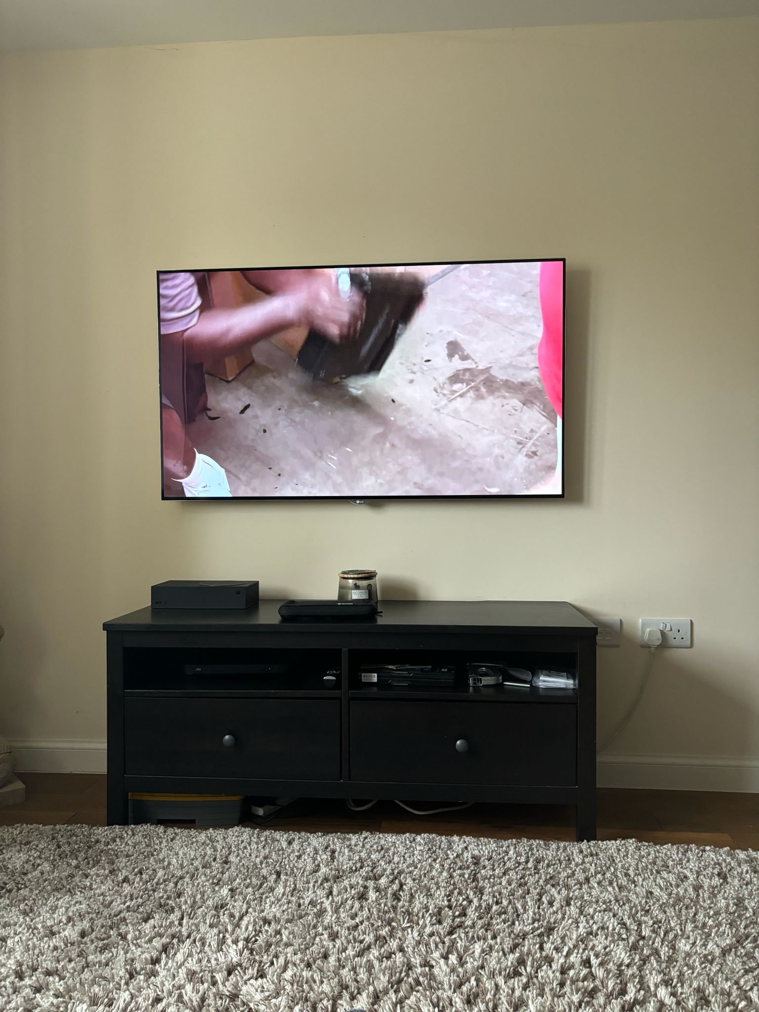 Living room with a mounted flat-screen TV on a wall, black TV stand underneath with electronic devices, and a beige shaggy rug in the foreground.
