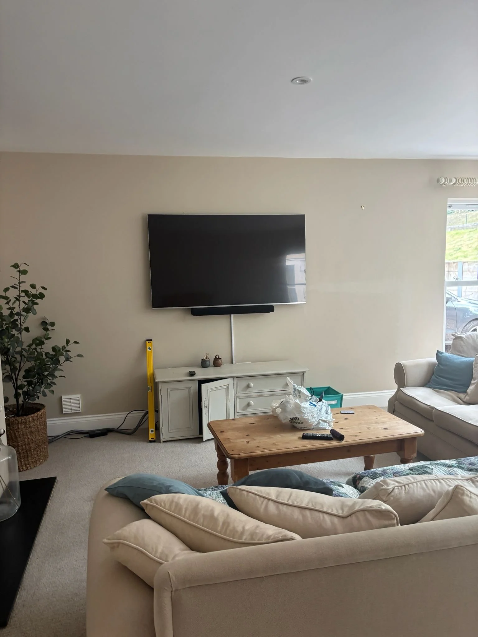 Living room with beige sofas, a wooden coffee table, a white cabinet, a mounted flat-screen TV, a potted plant, and various small items on the table and cabinet.