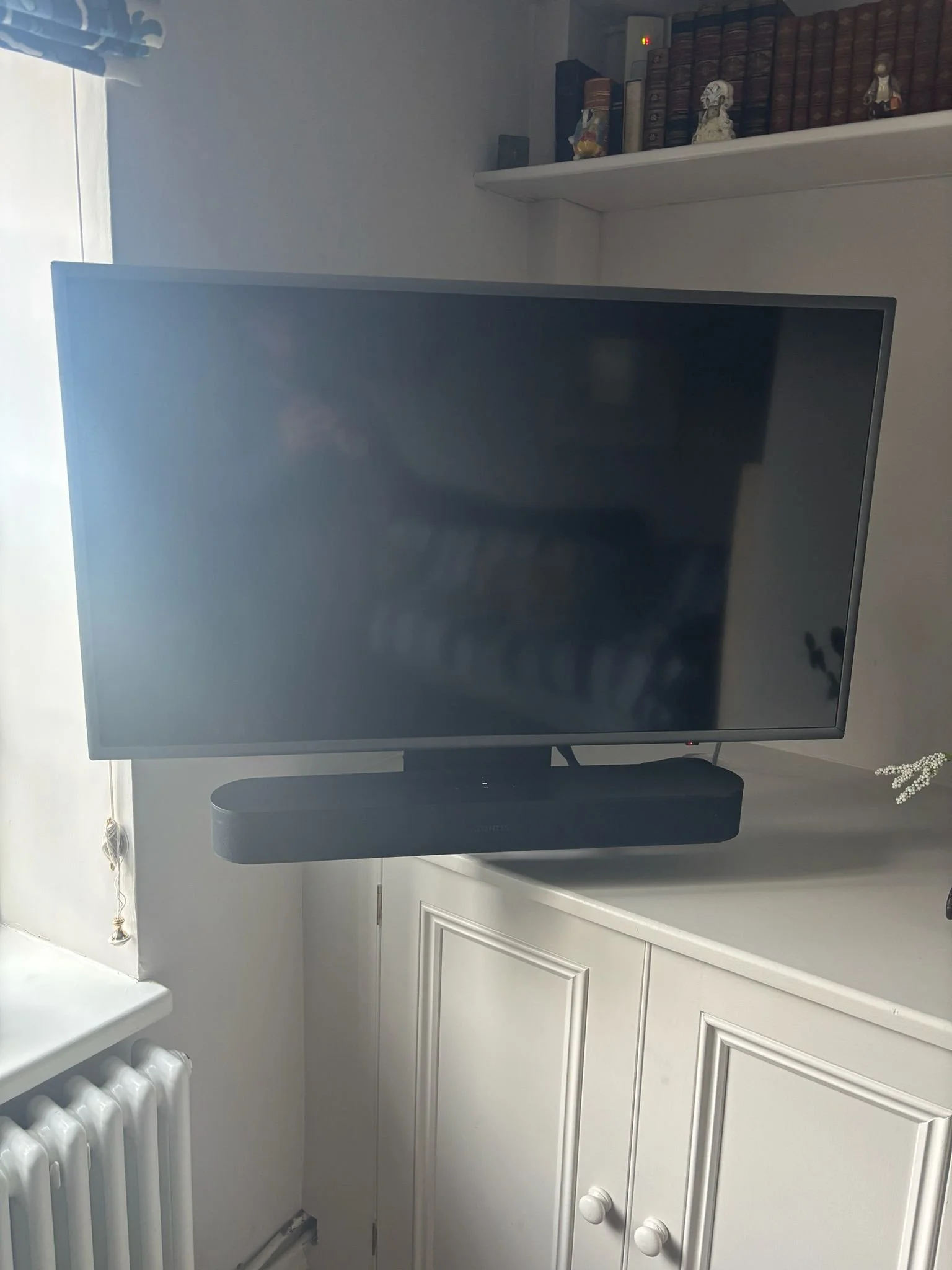 A large flat-screen television on a white cabinet, with a soundbar below it, in a room with a bookshelf containing books and decorative items in the background.