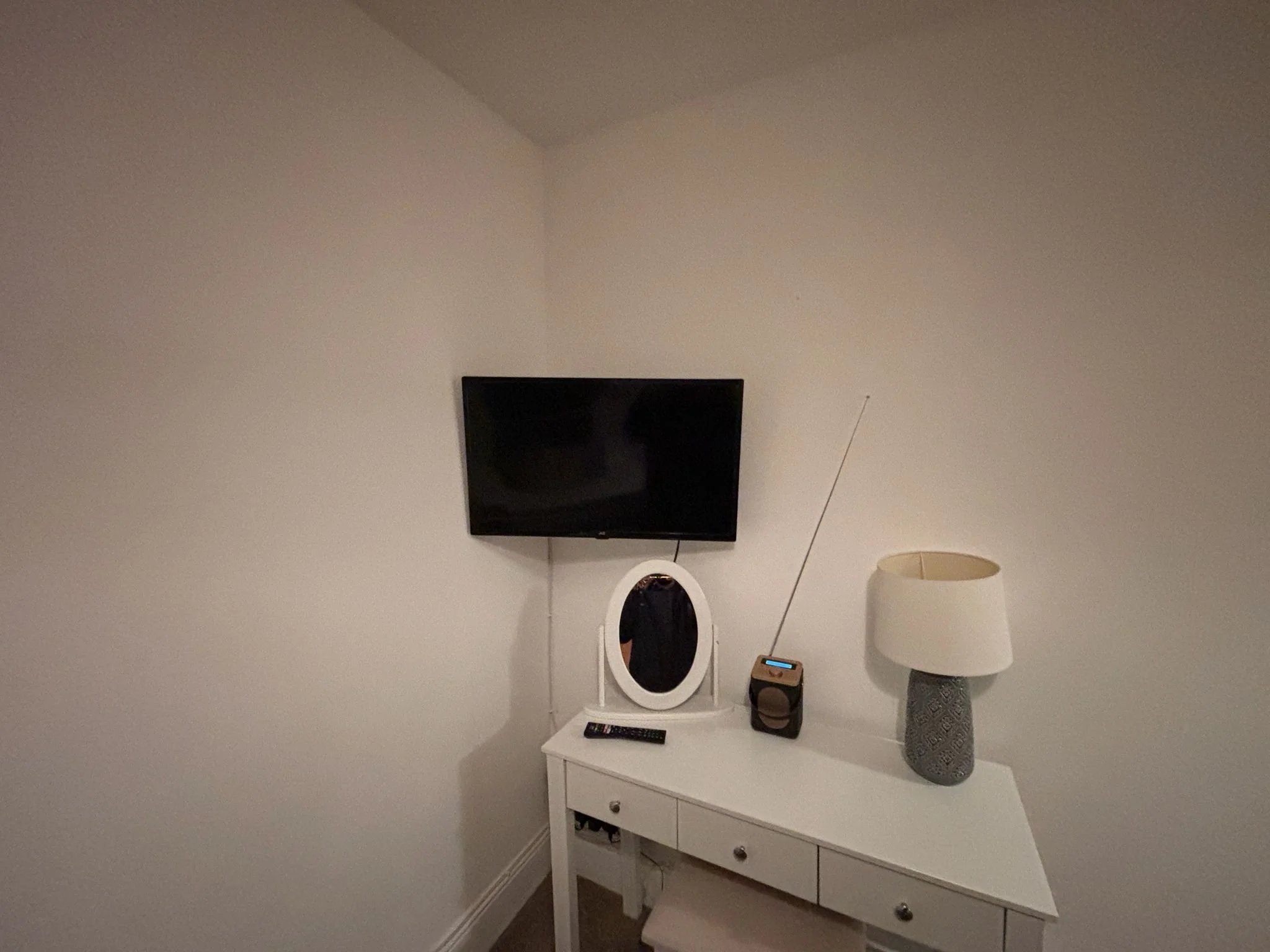 A corner of a room with a white dresser, a mirror, a remote, a radio with an antenna, a lamp with a textured gray base, and a mounted black flat-screen TV on a white wall.