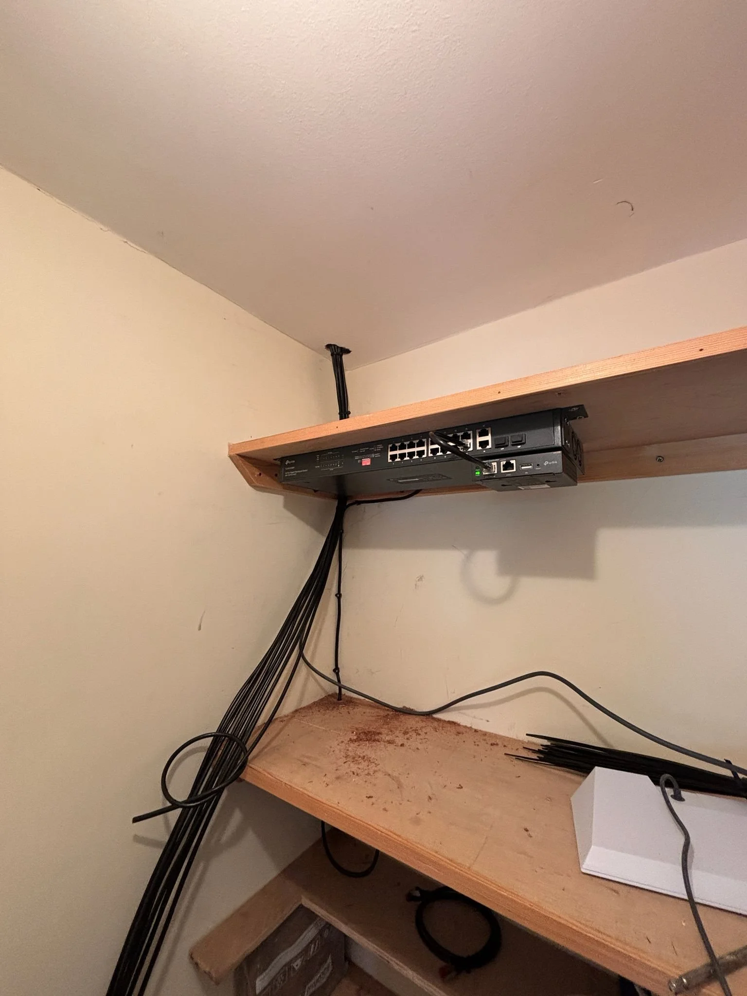 A network router mounted under a wooden shelf, with multiple black cables hanging down, some bundled together. The shelf is attached to a wall, and there is a second, lower wooden shelf with some dirt or debris and a white box on it.