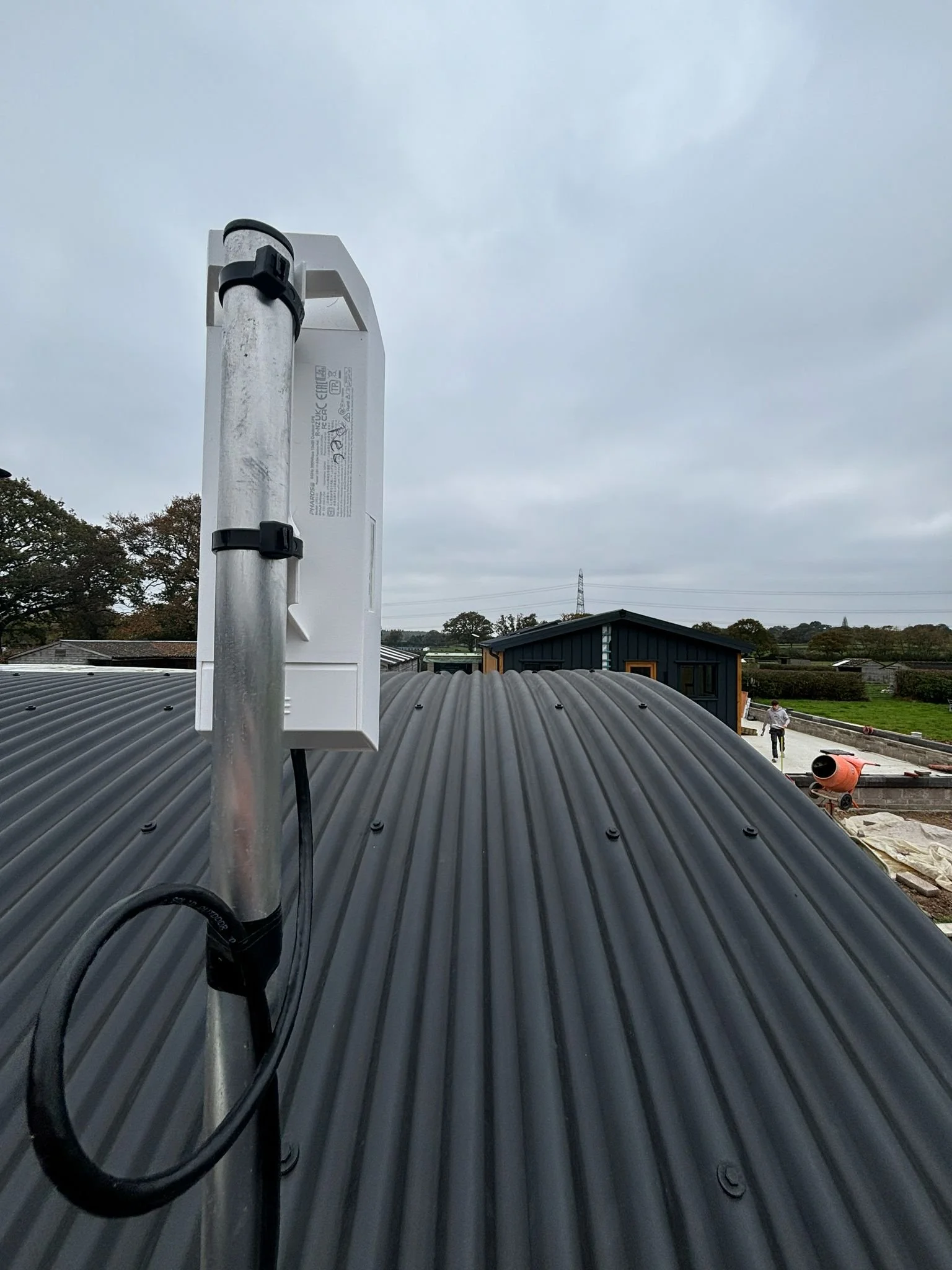 A close-up of a small antenna or signal device mounted on a metal pole on a black corrugated roof, with a cloudy sky and a rural landscape in the background.