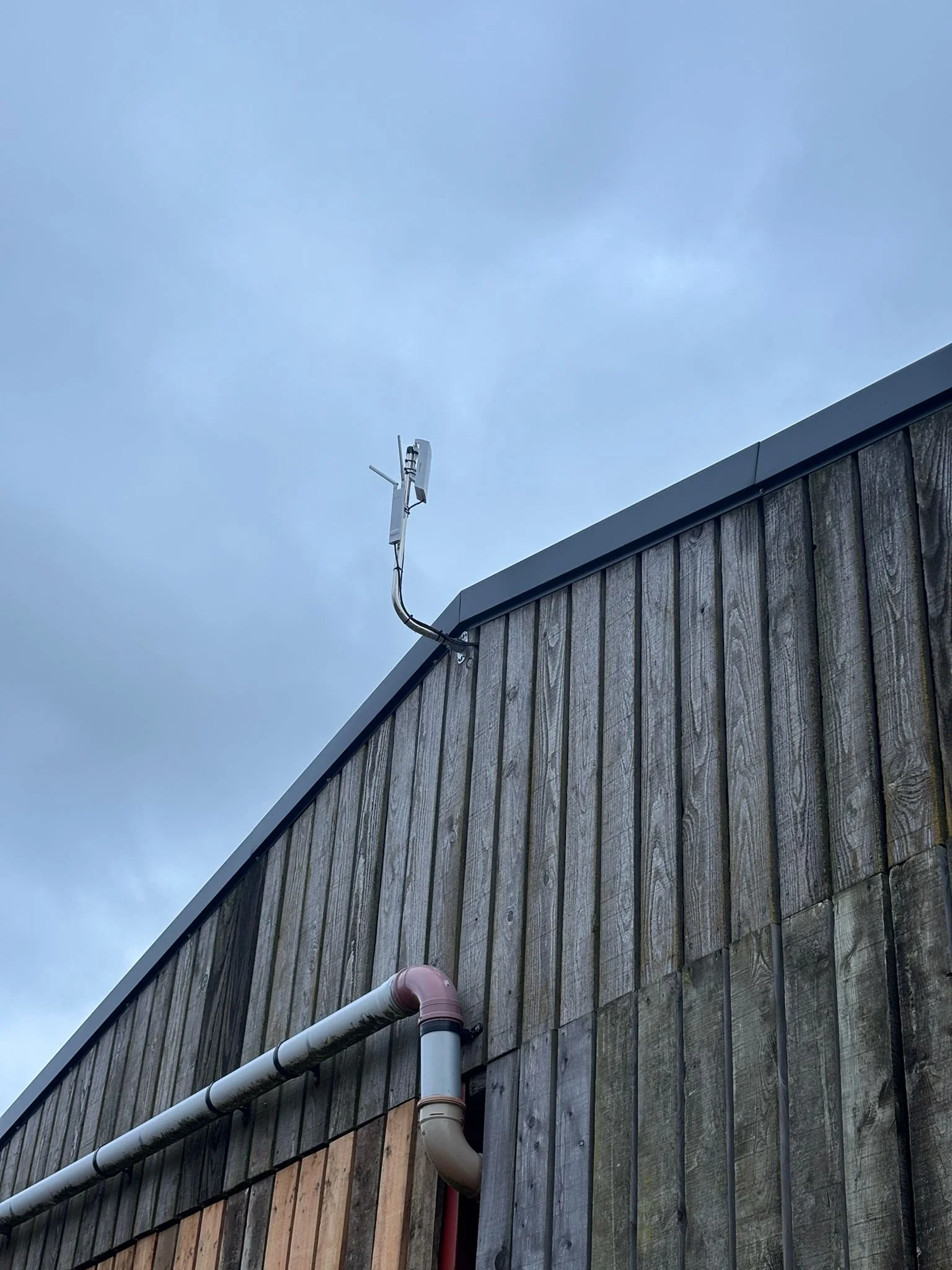 View of the exterior of a wooden building with a weathered look, featuring a white and pink pipe running along the side. A rooftop antenna is mounted at the top corner, with electrical wires connected to it. The background shows a cloudy sky.
