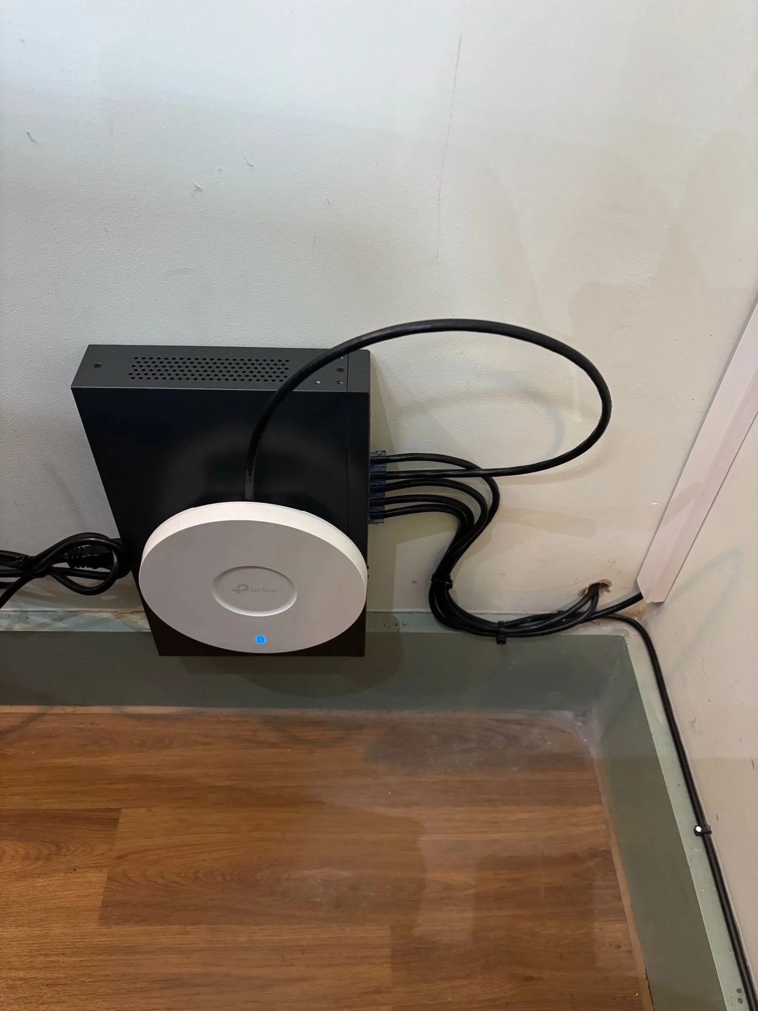 A TP-Link mesh Wi-Fi router sits on a black square device, connected to a wall and power outlet with multiple cables, on a wooden floor beside a white wall and baseboard.