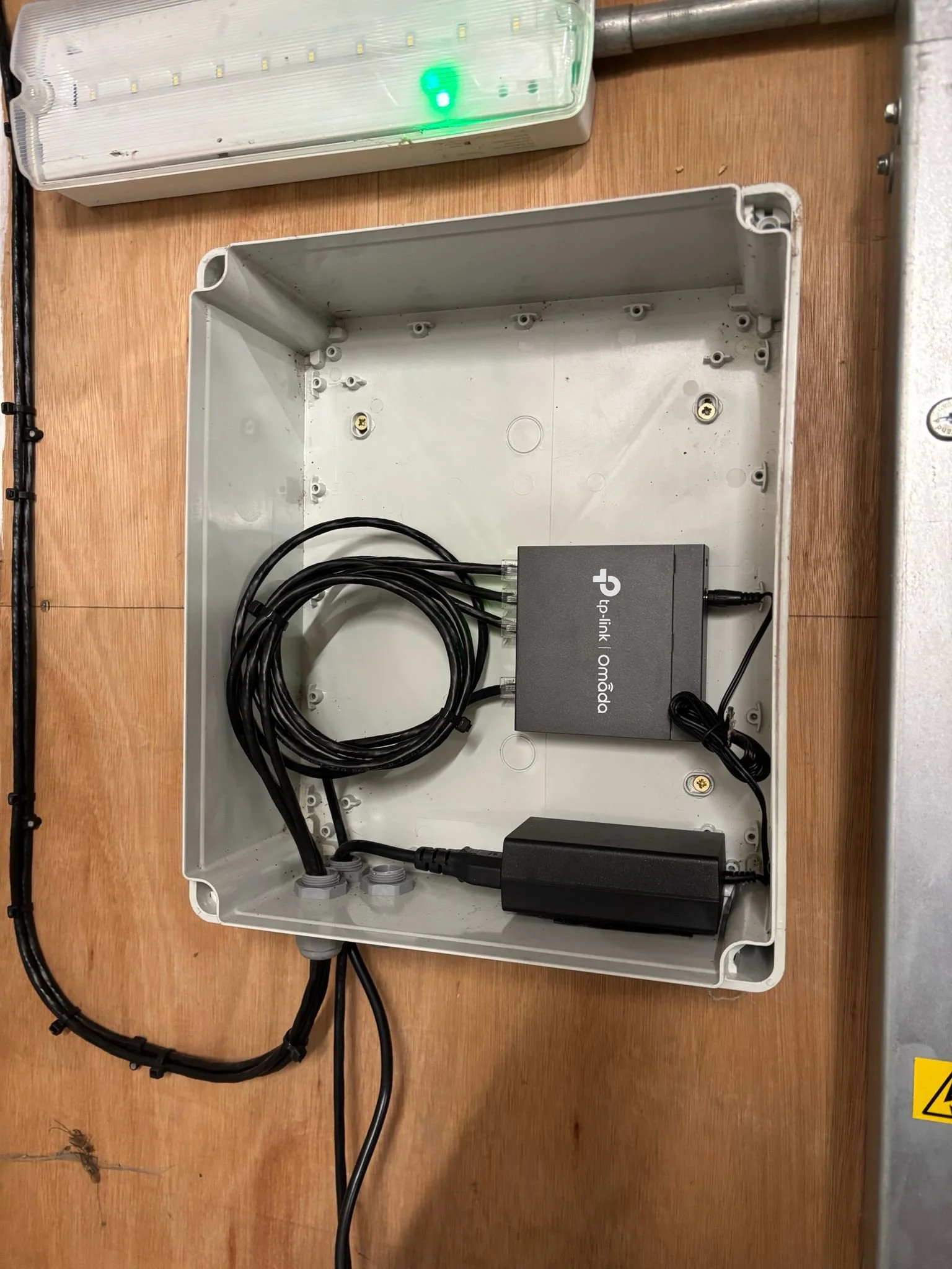 Open electrical box with connected black power supplies and a TPU-Link Omada device inside, placed on a wooden surface.