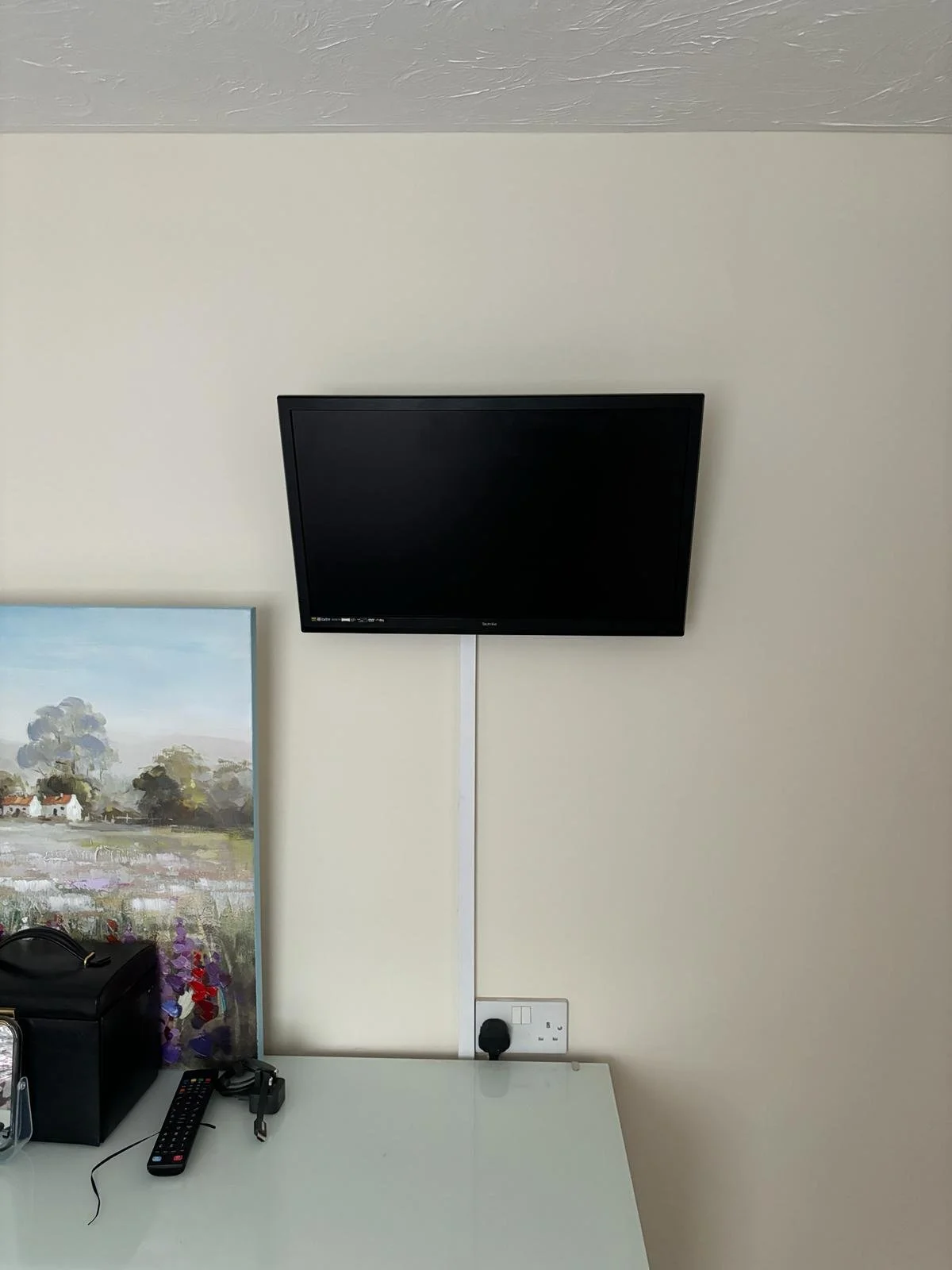 A wall-mounted flat-screen TV above a white table with a black device, a remote control, keys, and a power outlet.