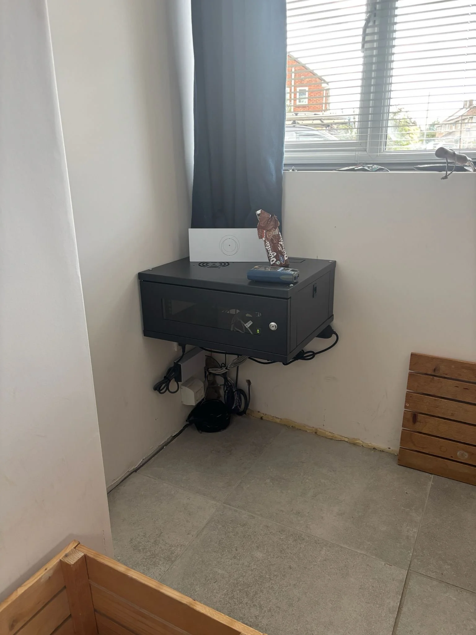 A black small cabinet with a glass door mounted on the wall in a room corner near a window. There are some objects on top of the cabinet, including a partially eaten snack, a remote control, and a white board or paper with a drawing. Electrical cords