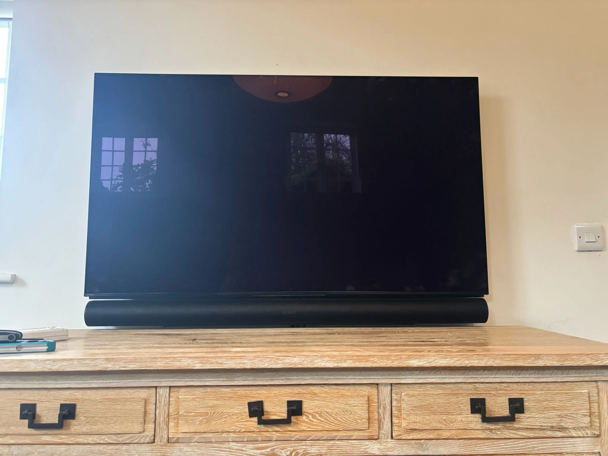 A large flat-screen television on a wooden dresser with a soundbar below it, and a wall with a window reflecting trees and sky behind it.