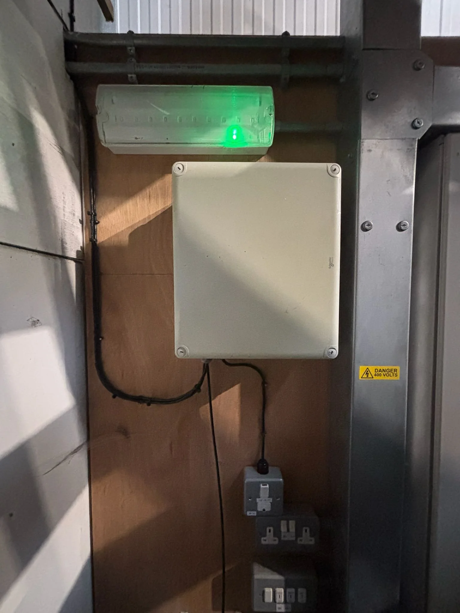 Electrical panel with a white box, a green-lit indicator light, and multiple power outlets connected to a wall and metal structure.