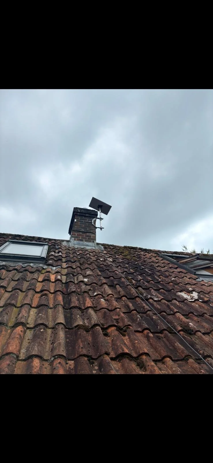 Rooftop with weathered clay tiles, a chimney, a skylight window, and a Starlink dish installed on the chimney against a cloudy sky.