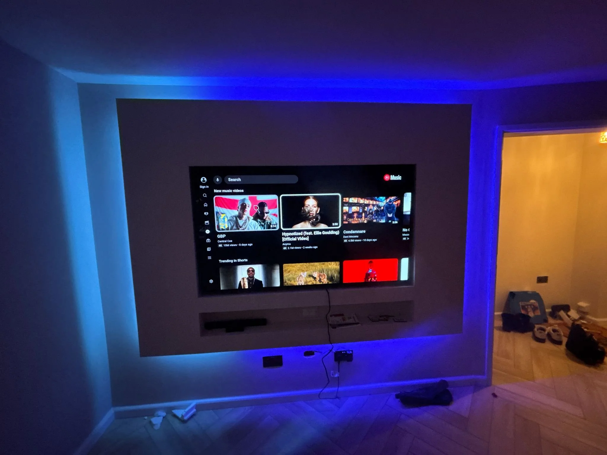 A living room with a wall-mounted television displaying a music app, illuminated with blue LED strip lights. There are various objects on the floor and a doorway to another room illuminated with warm yellow lighting.