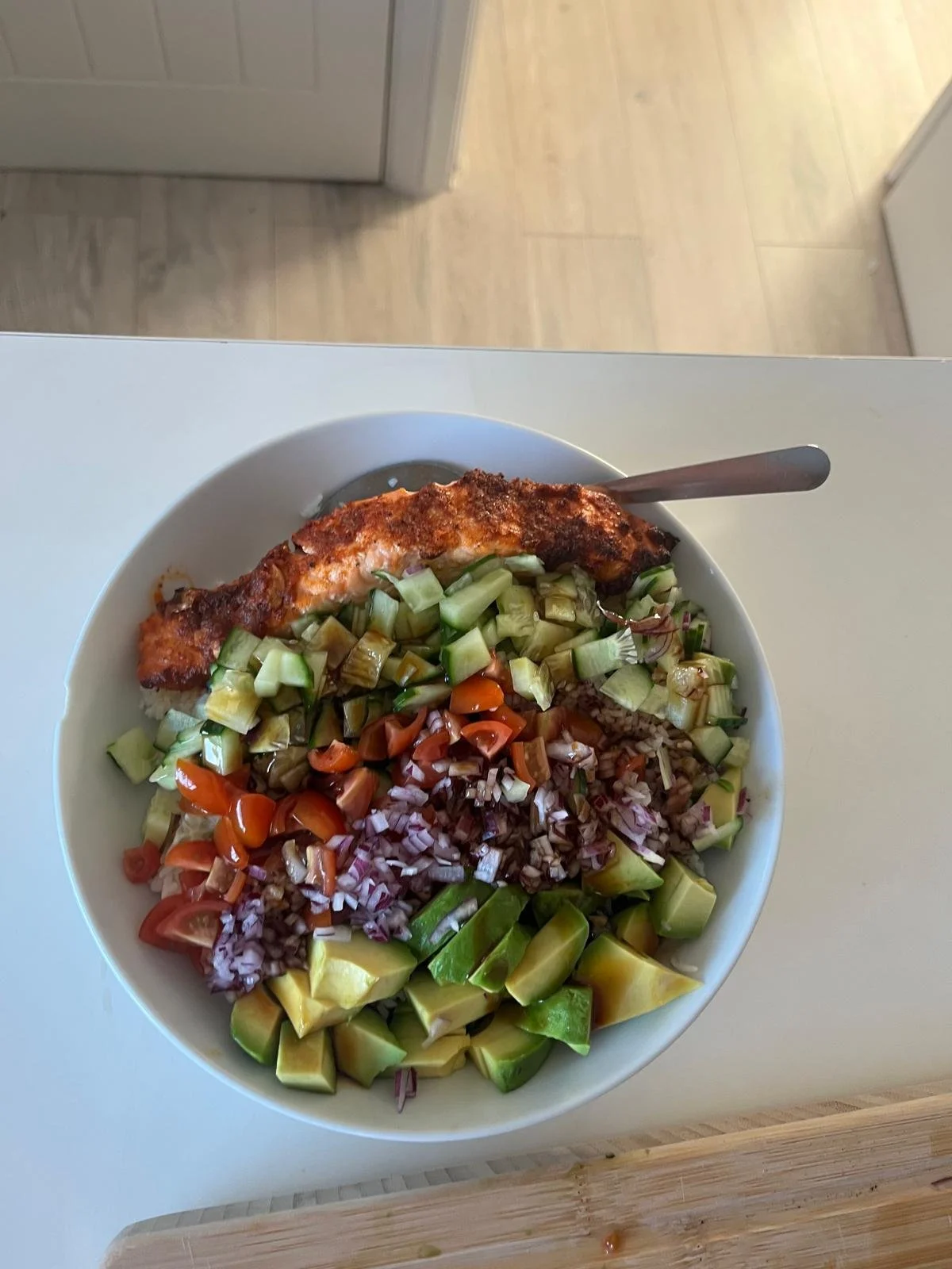 Bowl of rice topped with baked salmon, chopped cucumbers, cherry tomatoes, red onions, and sliced avocado on a white table.