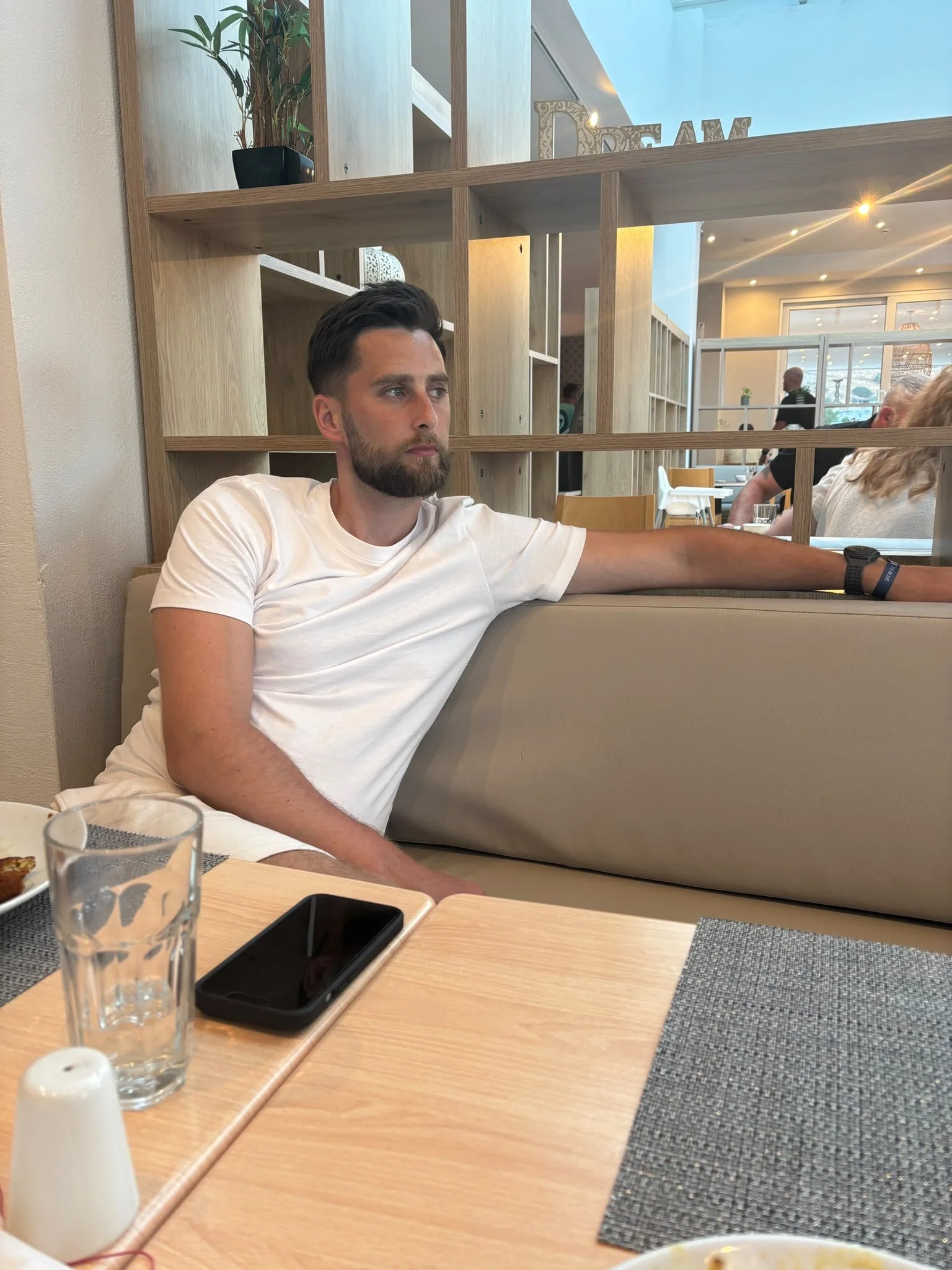 A young man with dark hair and a beard sitting at a wooden table in a restaurant or cafe, with his arm resting on a beige cushioned seat. He wears a white t-shirt and a black wristwatch. On the table are a glass of water, a smartphone, a small salt shaker, and part of a plate with food. The background shows a wooden shelf with decorative items, a large window with natural light, and other diners.