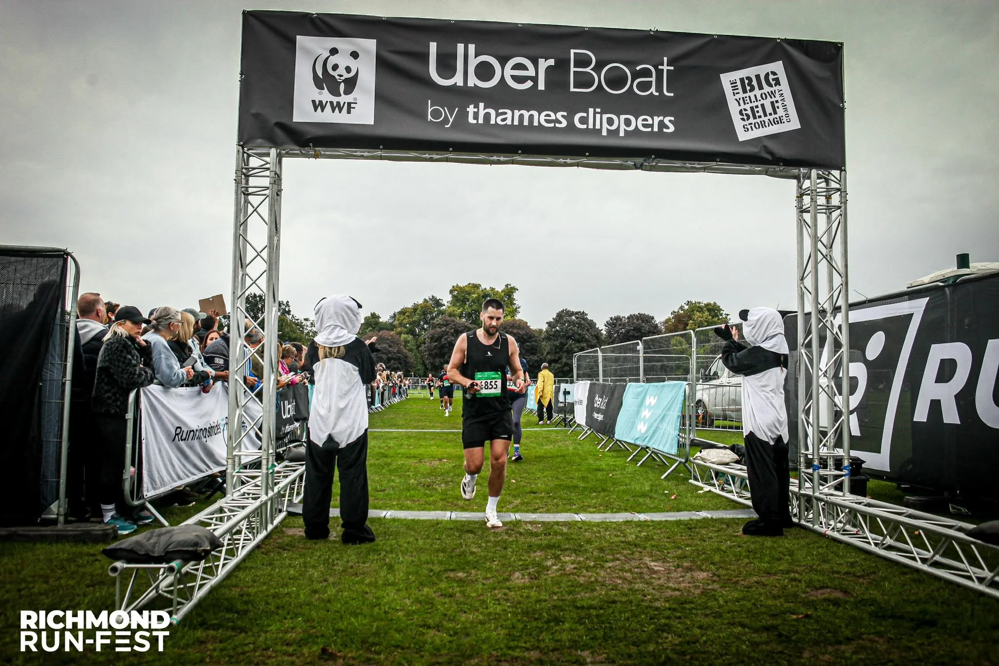 A male runner with bib number 855 finishing a race at the Richmond Richmond Run Fest, passing through a finish line under a black banner that reads Uber Boat by Thames Clippers and WWF. Spectators are on either side of the finish area, some taking photos.