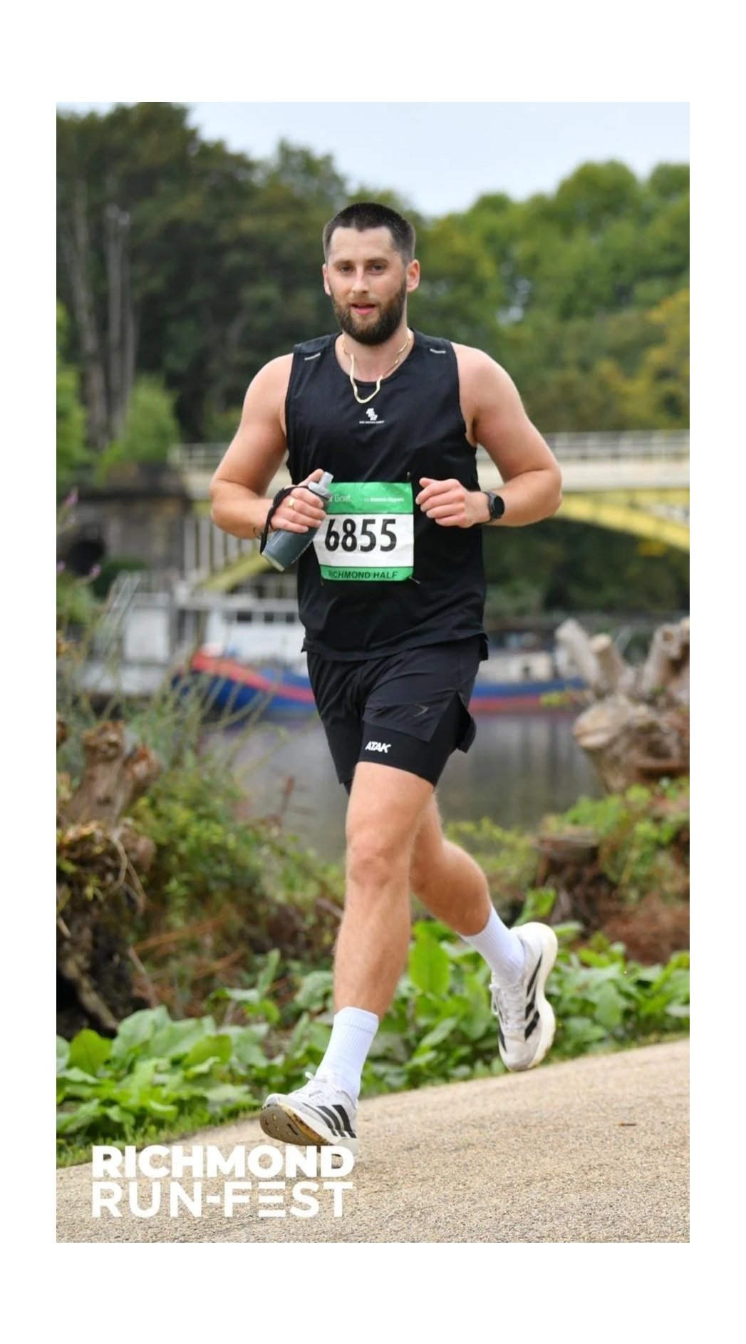 A male runner dressed in black athletic wear with a race bib number 6855 running outdoors near a river, with trees and a bridge in the background, during the Richmond Run Fest.