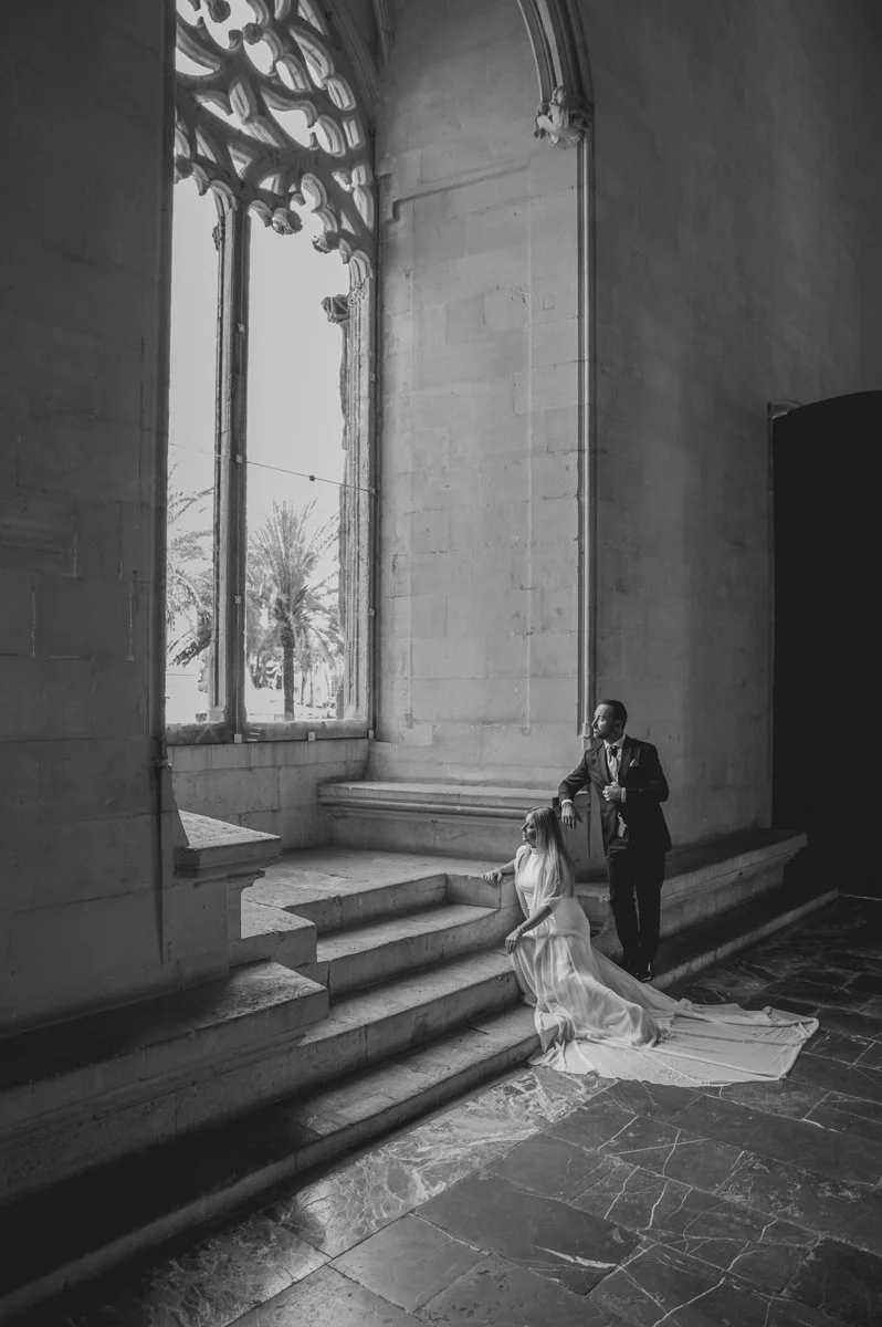 Pareja en un interior de iglesia o capilla, con un gran ventanal y decoraciones góticas; la novia con vestido largo y el novio en traje oscuro, en una escena que parece ser de una boda.