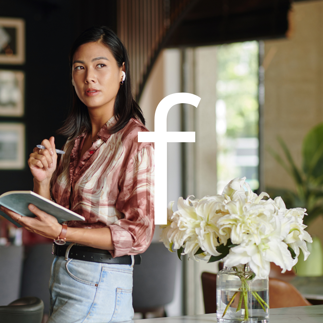 A woman standing in a modern interior with natural light, holding a notebook and pen, wearing earphones and a satin blouse, with a bouquet of white lilies on a table nearby.