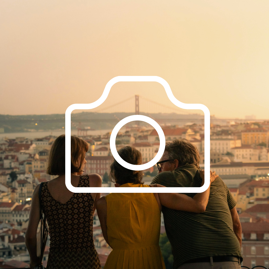 Three people sit together on a balcony, overlooking a cityscape at sunset with a bridge in the background.