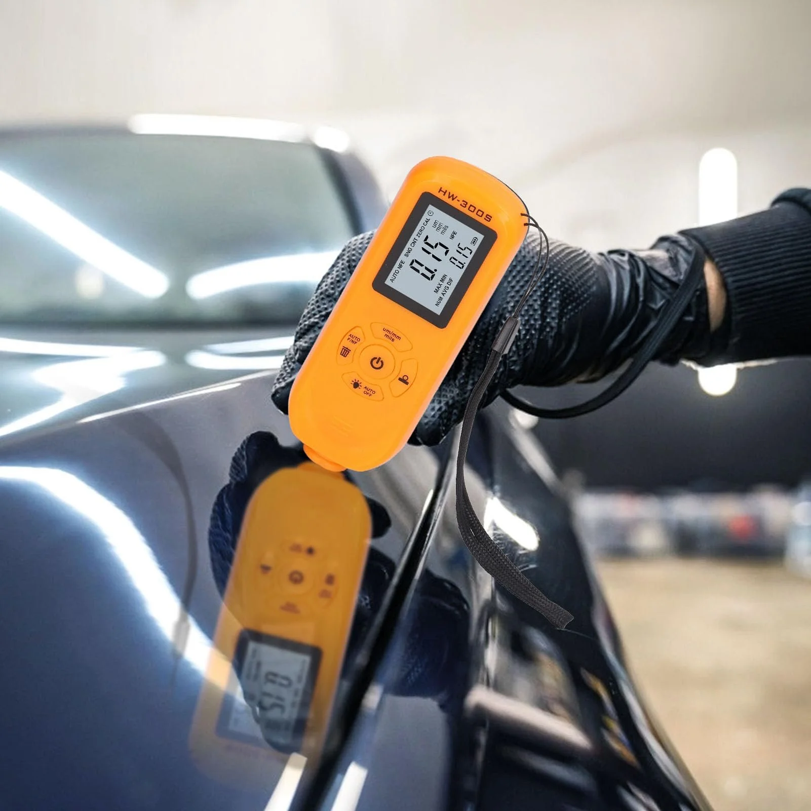 A person wearing black gloves is measuring the paint thickness on a dark-colored car's surface using an orange digital paint gauge, with the car's reflective surface showing the measurement device above.