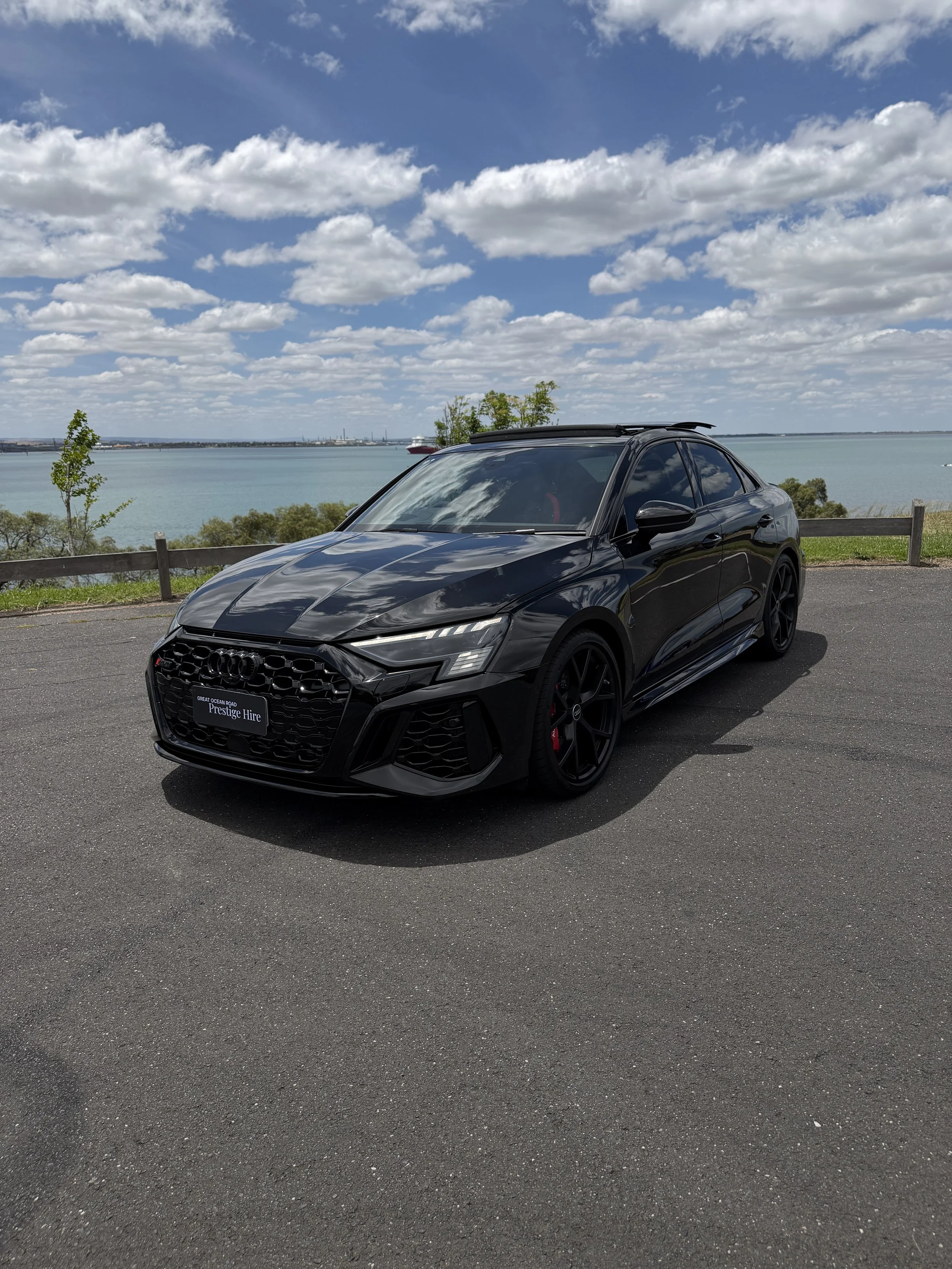 A black Audi sedan parked on an asphalt road near a body of water with trees and a cloudy sky in the background.