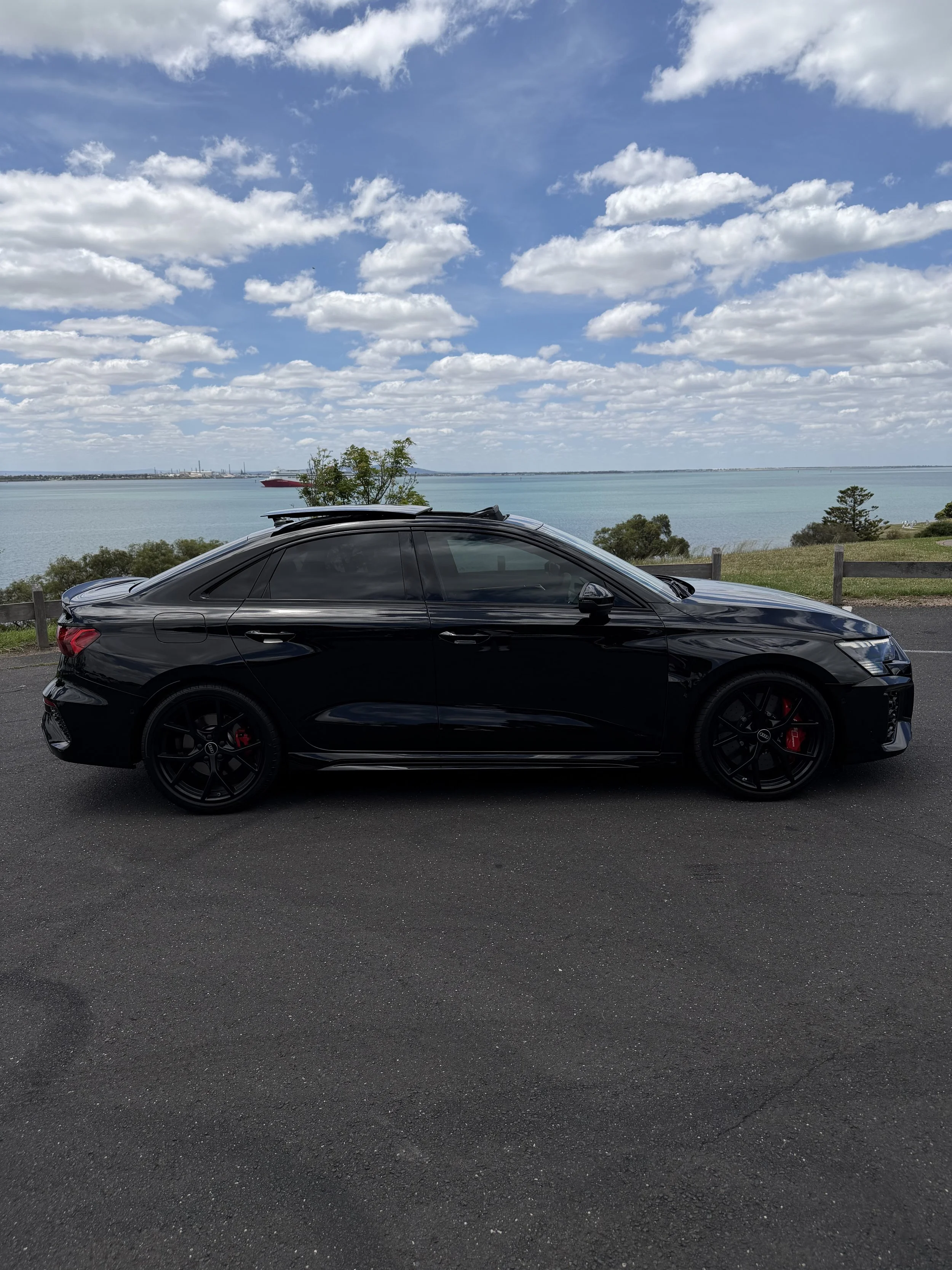 Black luxury sedan car parked on roadside with lake and cloudy sky in background.