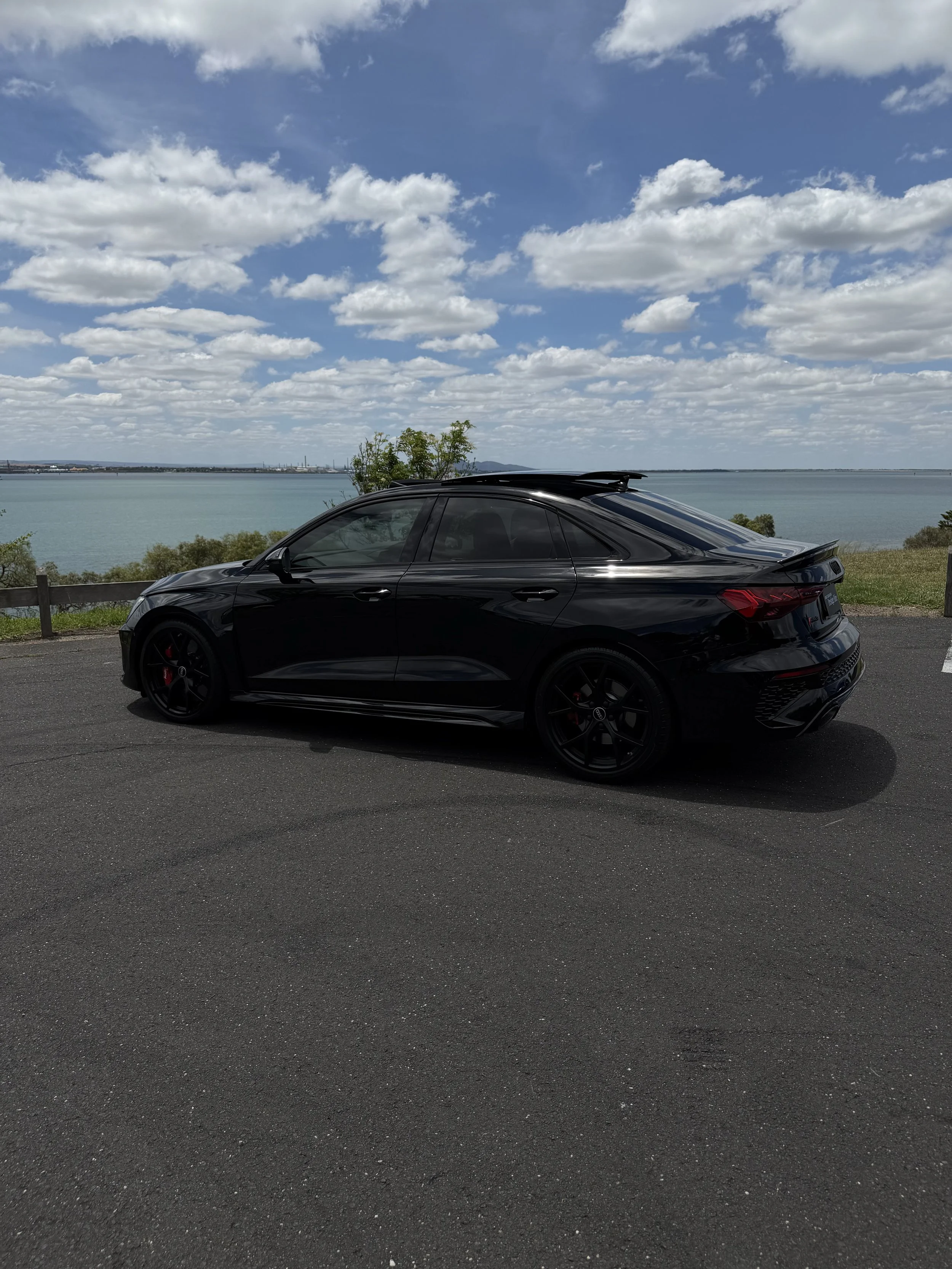 Black sedan car parked on the side of a road with a lake and cloudy sky in the background.