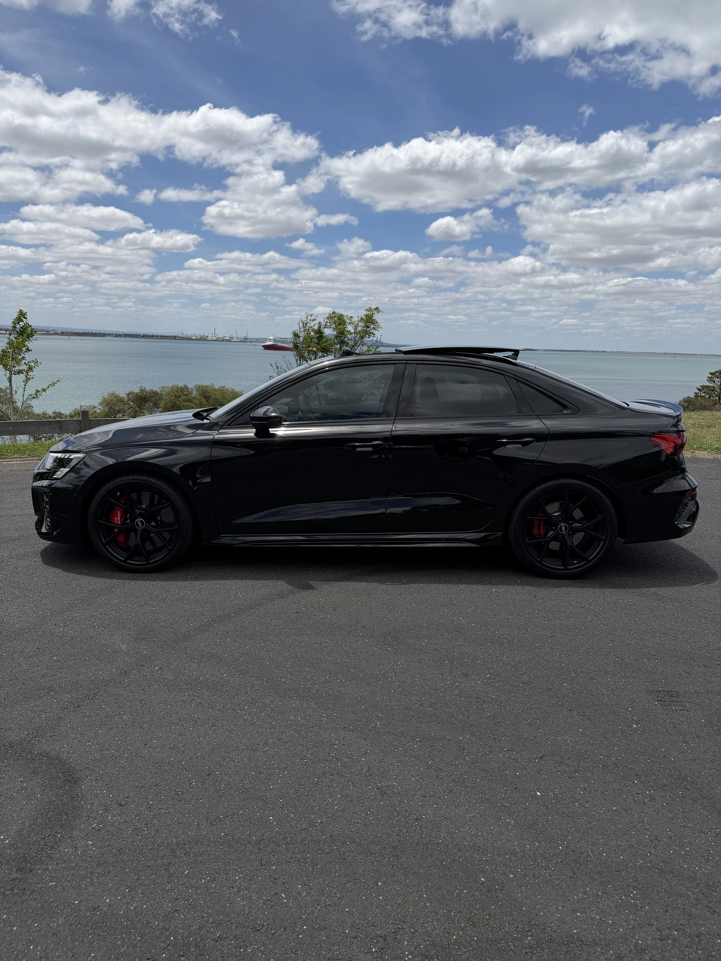A black coupe car parked on the side of a road with a lake, trees, and cloudy sky in the background.