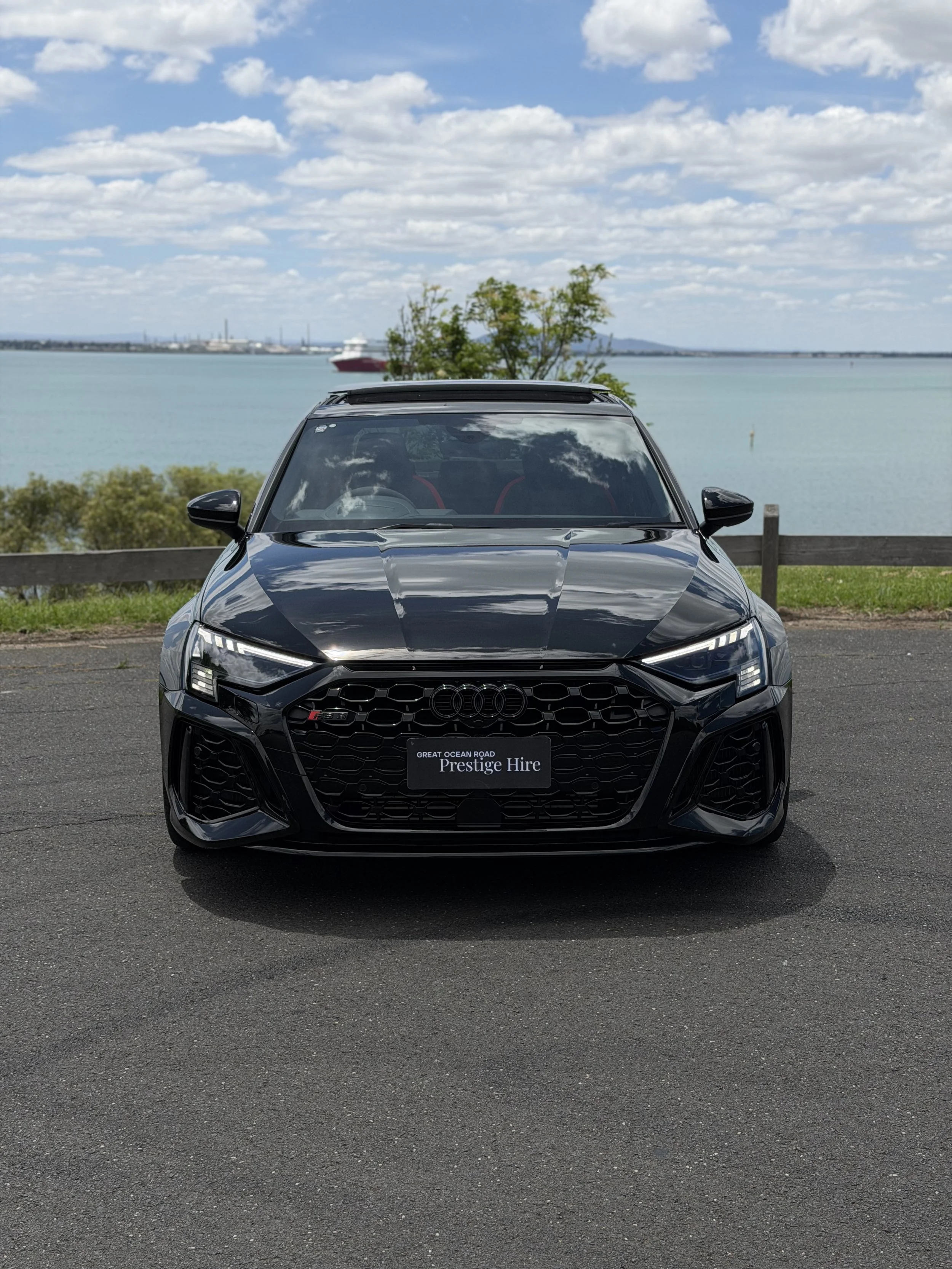 Black Audi car parked on pavement near water, with sky and clouds in background.