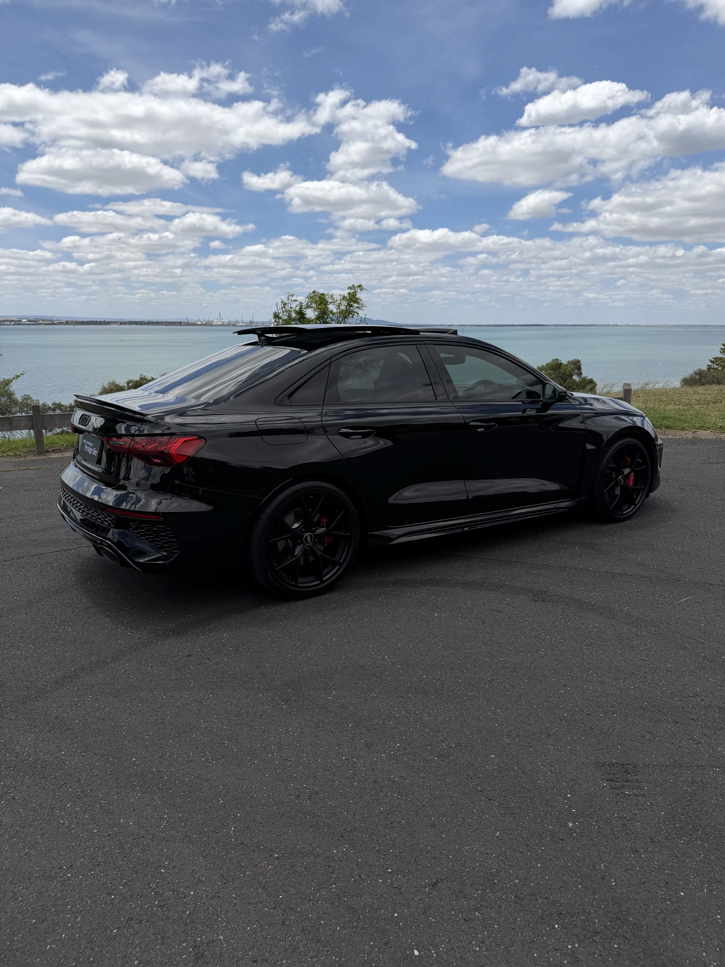 Black hatchback car parked on a paved road beside a body of water with a cloudy sky overhead.