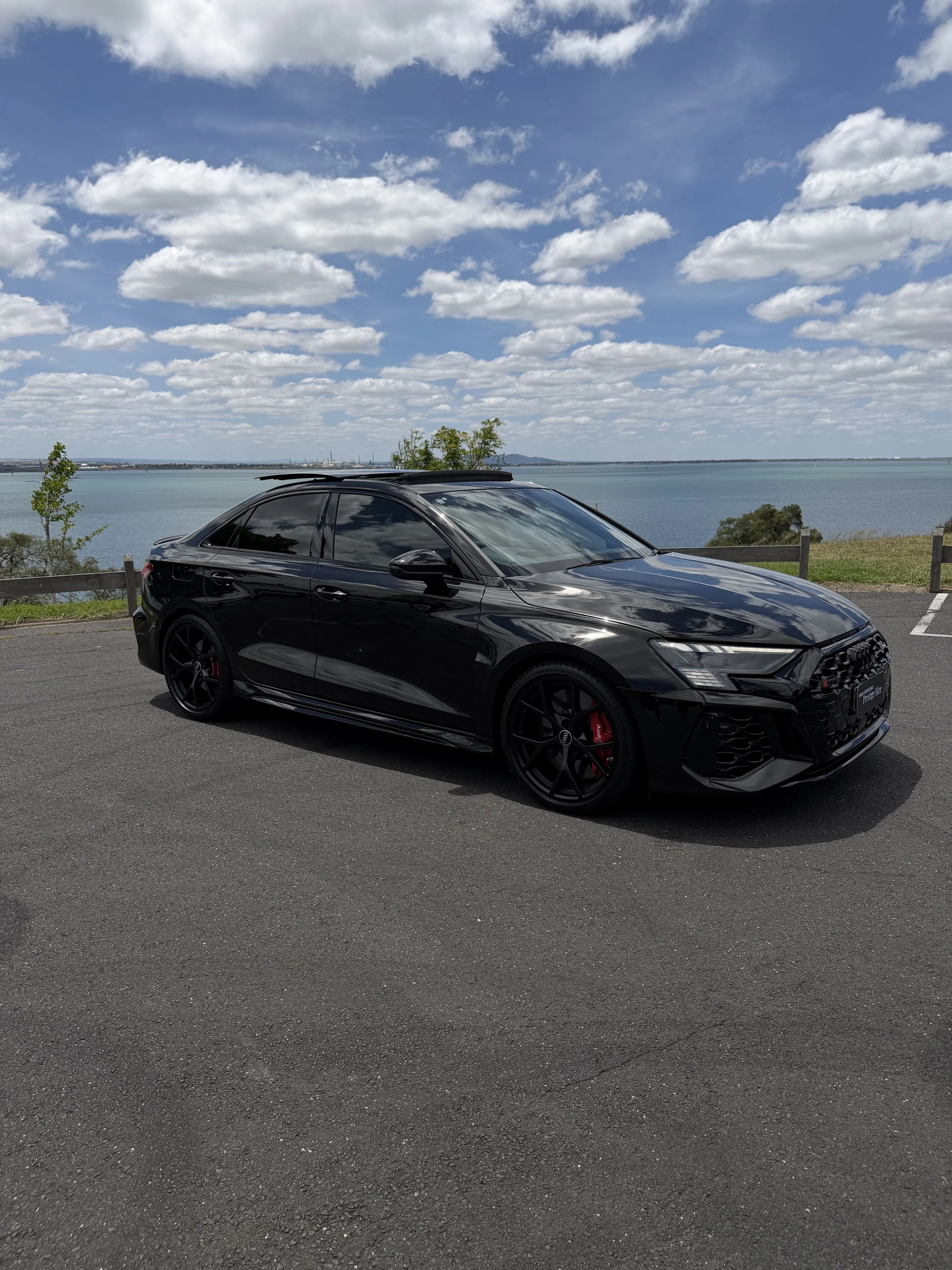 A black sports car parked on a paved lot near a body of water with a scenic view of the sky, clouds, and distant landscape.